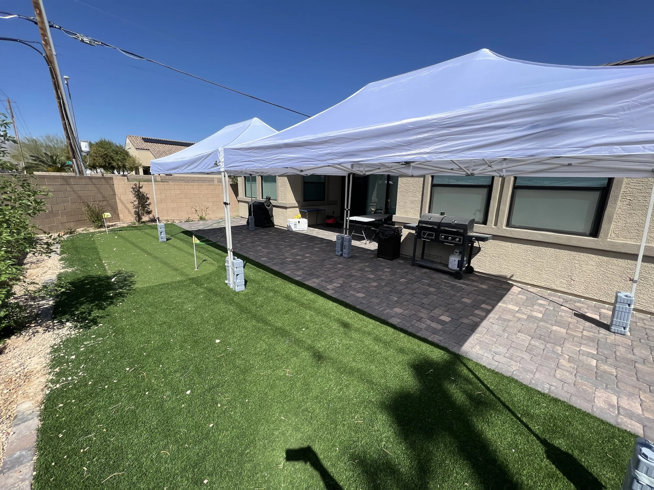 Backyard patio with artificial turf, a barbecue grill, and white canopy tents on a sunny day.