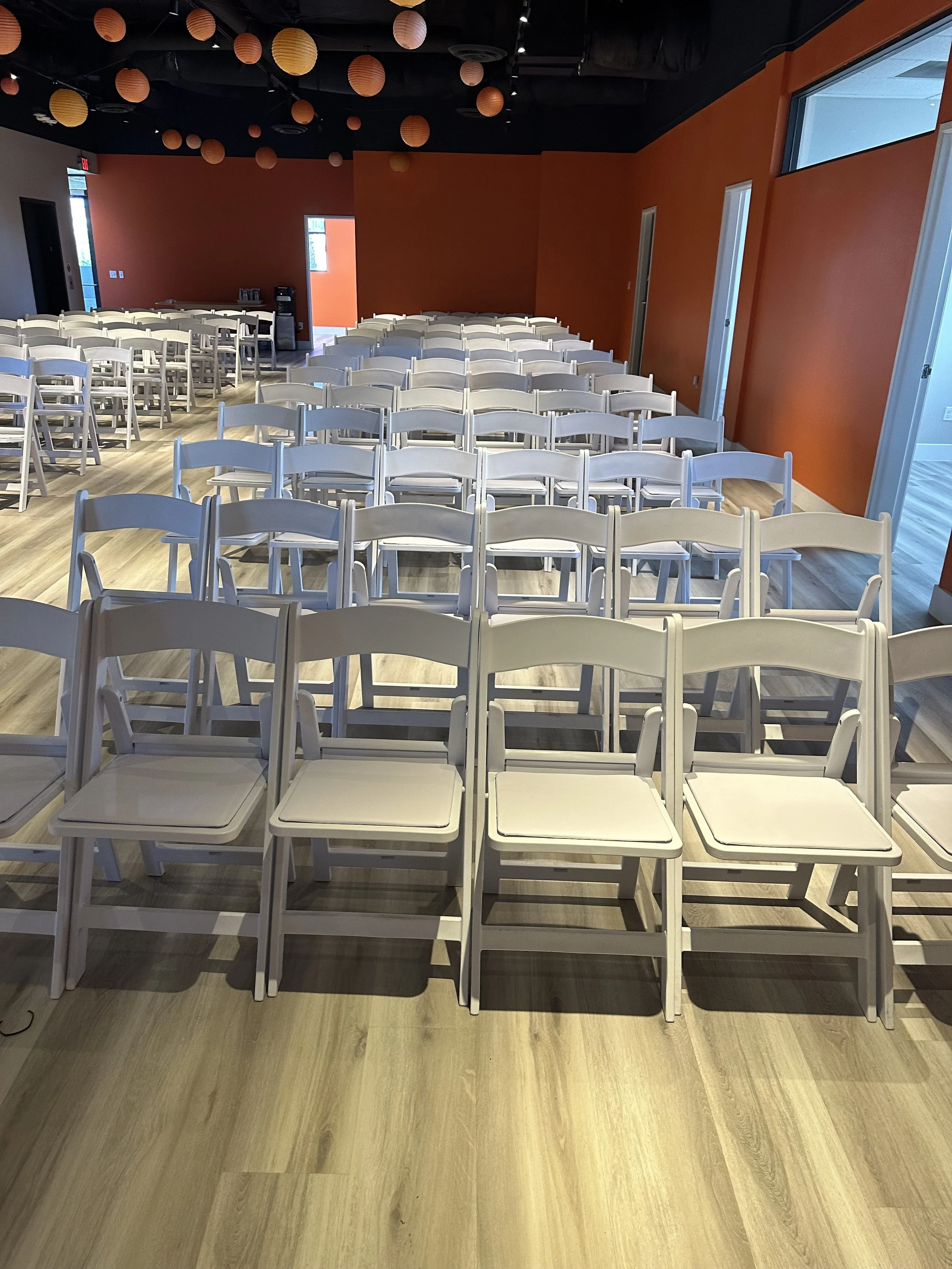 Rows of white folding chairs in a room with orange walls and hanging decorations.