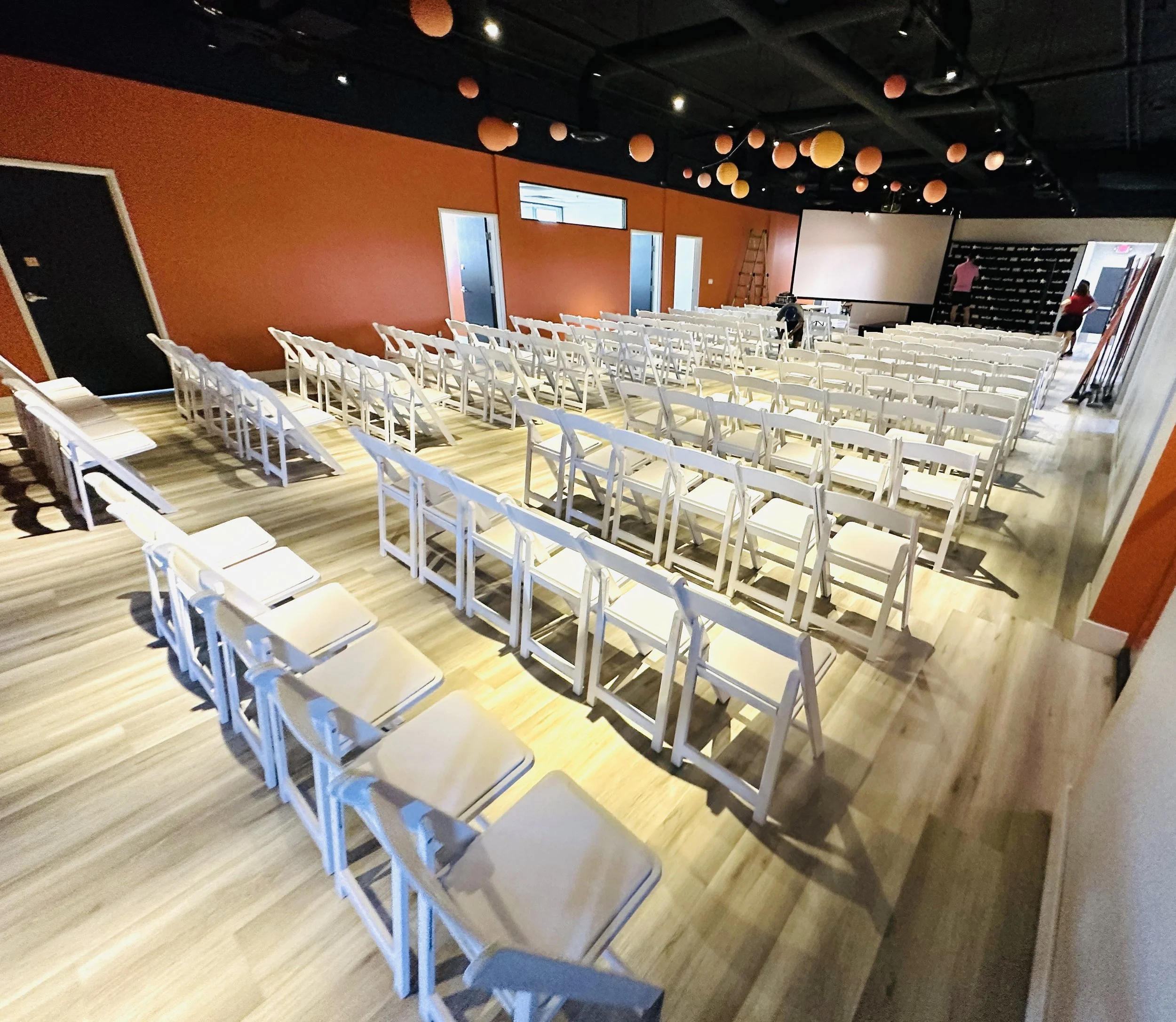 Empty event space with rows of white chairs, an orange wall, and a large projection screen.
