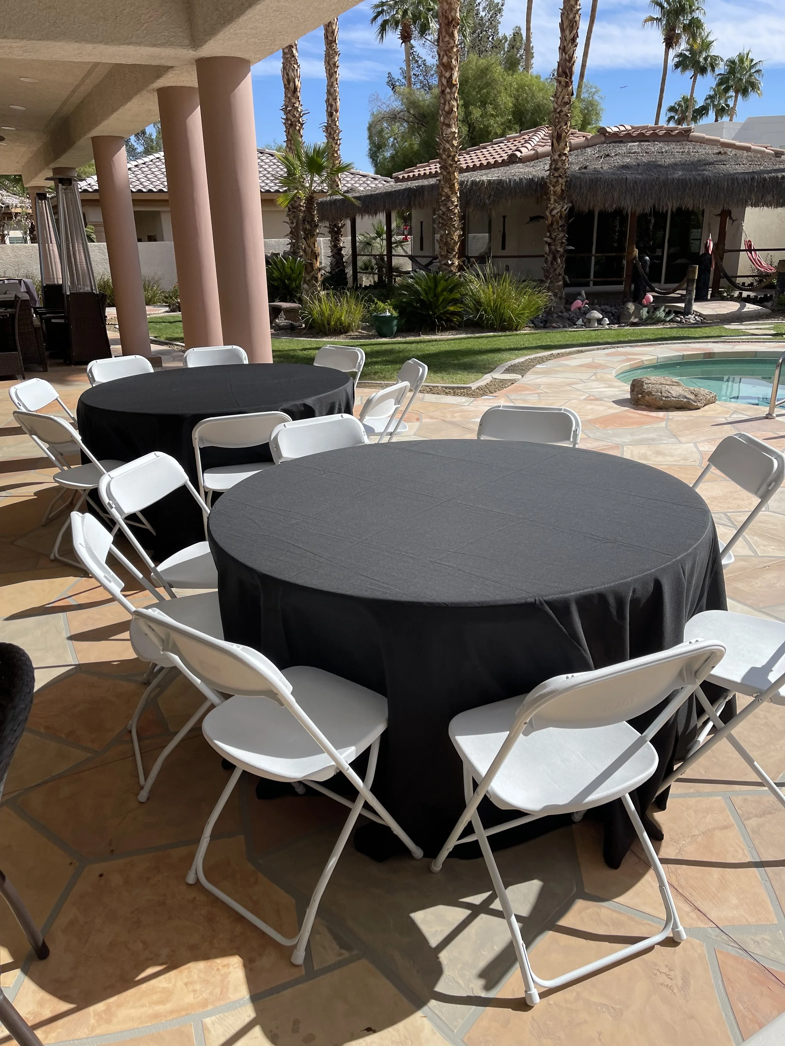 Outdoor patio with round tables covered in black tablecloths, surrounded by white folding chairs, beside a pool and garden with palm trees.