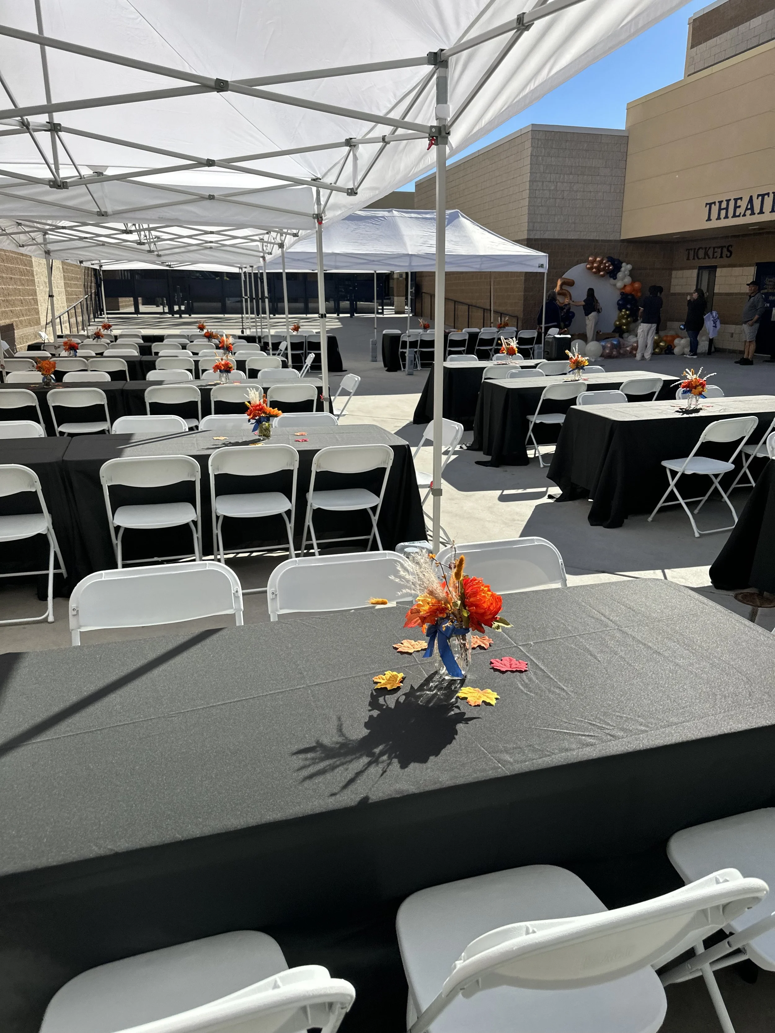 Outdoor event setup with tables and chairs under white tents, decorated with fall-themed centerpieces on black tablecloths, located near a theater entrance.