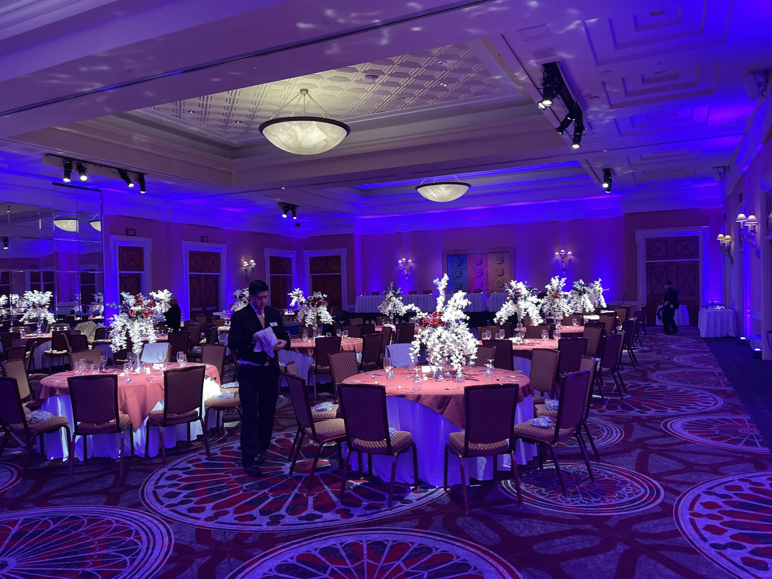 Elegantly decorated banquet hall with round tables, floral centerpieces, and purple lighting.