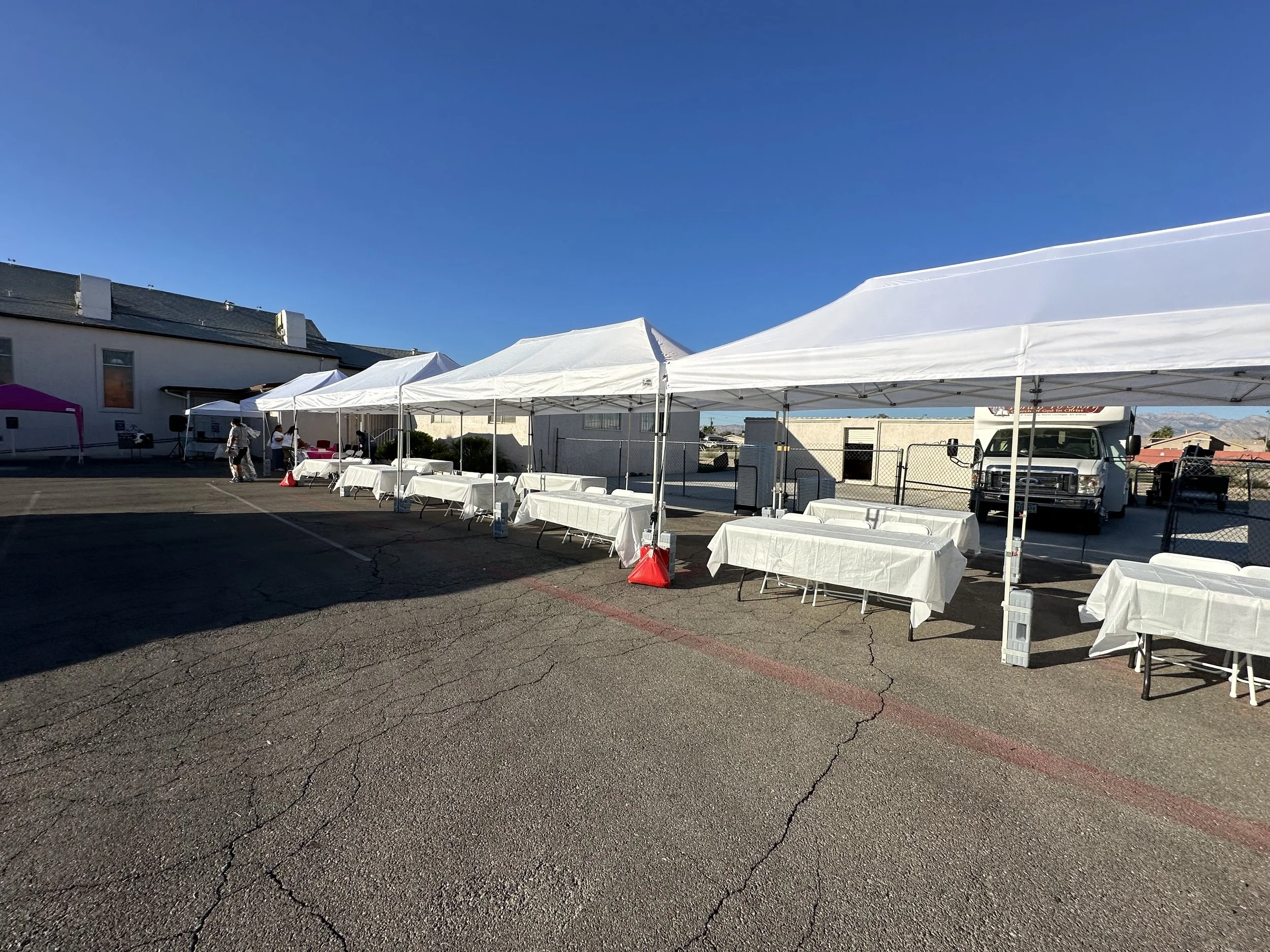 Outdoor event setup with white tents and tables covered in white cloths, on a paved surface, with a truck and building in the background under a clear blue sky.