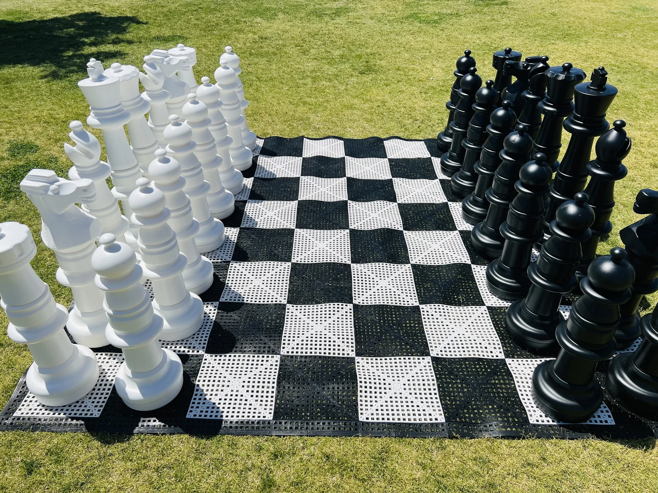 Large outdoor chess set with black and white pieces on a checkered mat, placed on grass.