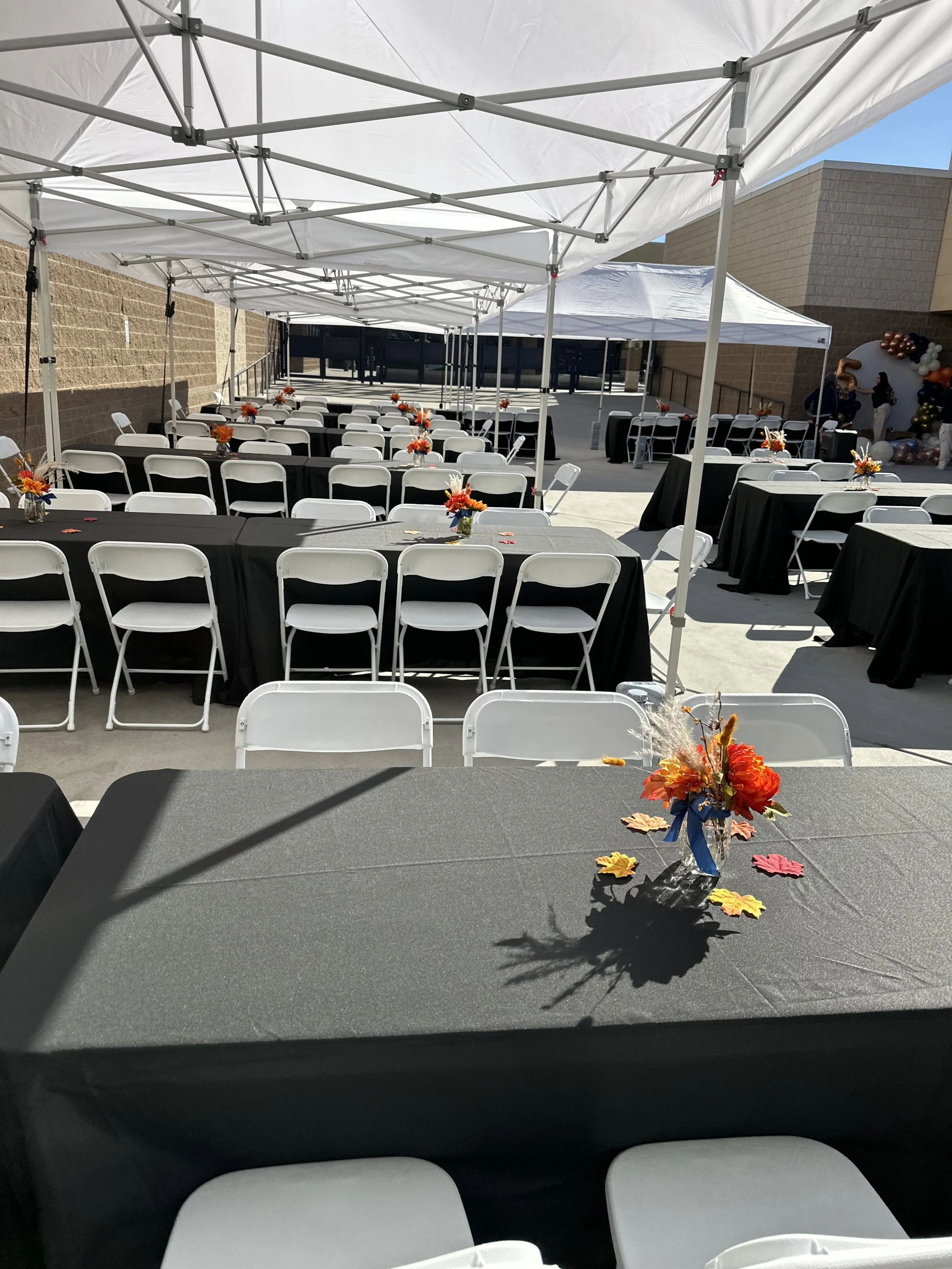 Outdoor event setup with white canopy tents, rows of tables covered with black tablecloths, white folding chairs, and floral centerpieces.