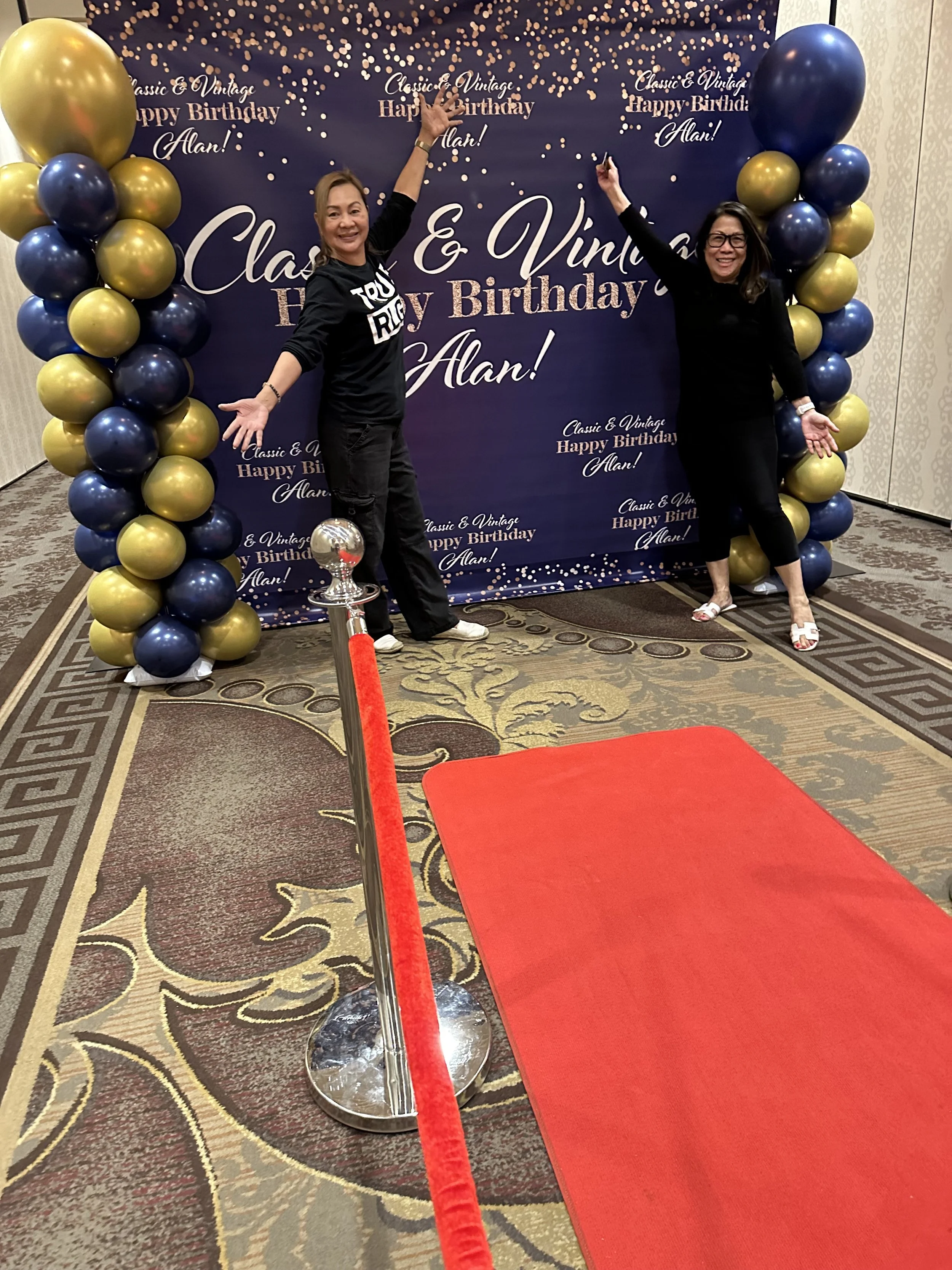 Two women posing in front of a birthday-themed backdrop with balloons and a red carpet path.