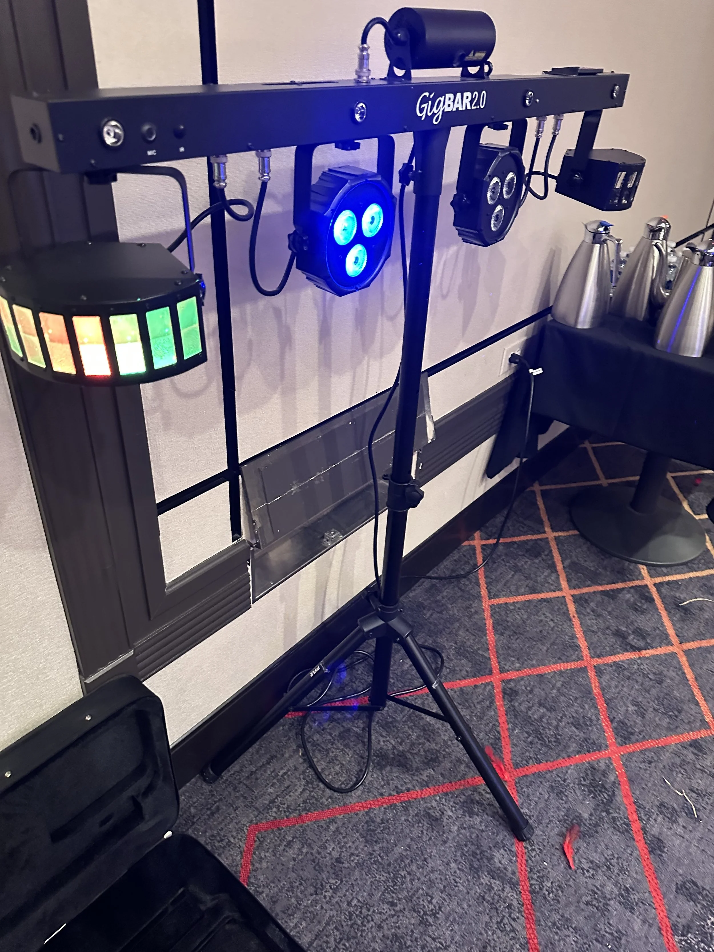 GigBAR 2.0 lighting system setup in a room, featuring multiple stage lights on a tripod stand, with a black case and metal pitchers nearby.