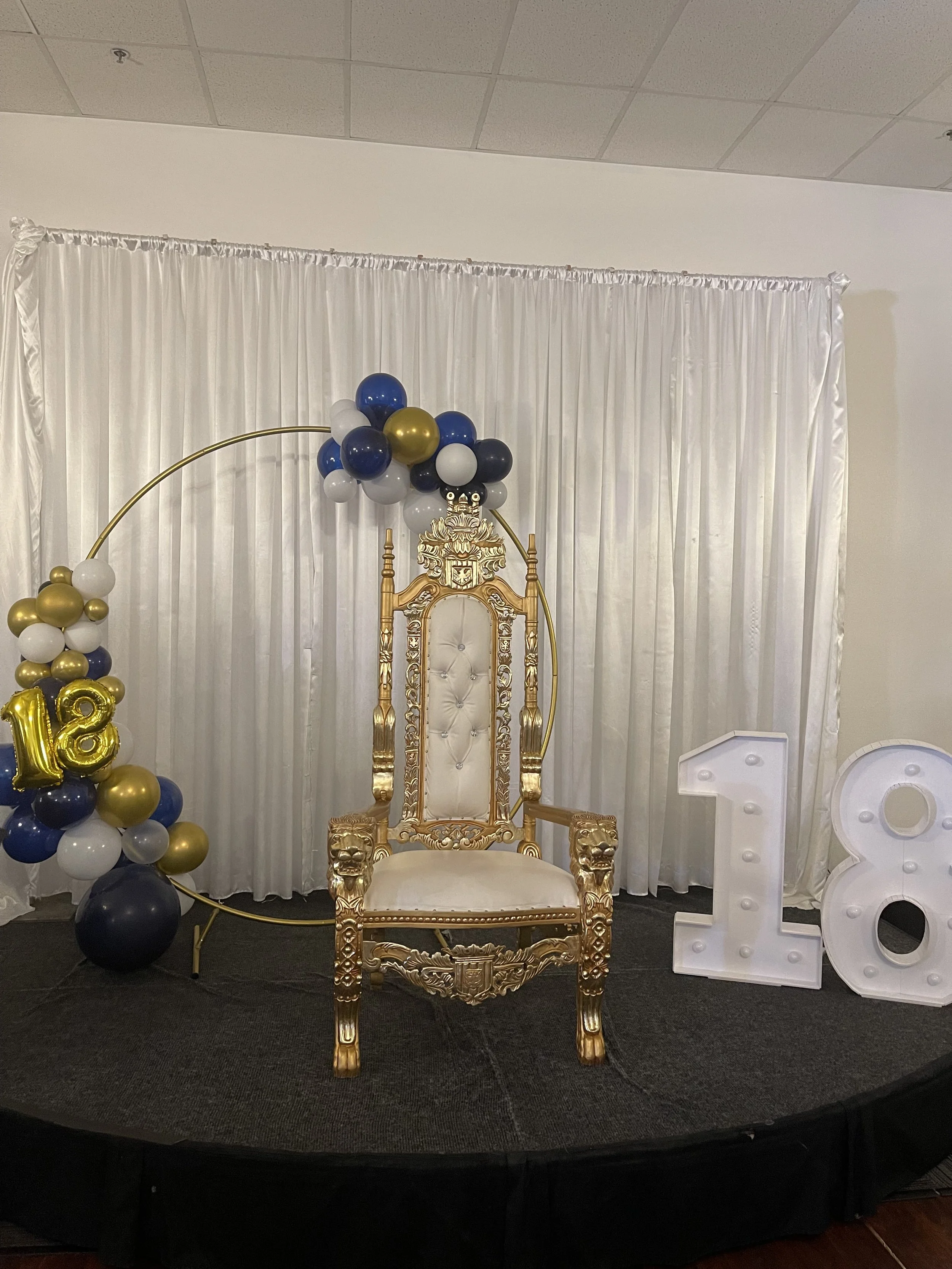 Elegant event setup with a gold throne chair, white backdrop, balloon arch in blue, white, and gold, and large illuminated '18' numerals. Ideal for 18th birthday celebration.