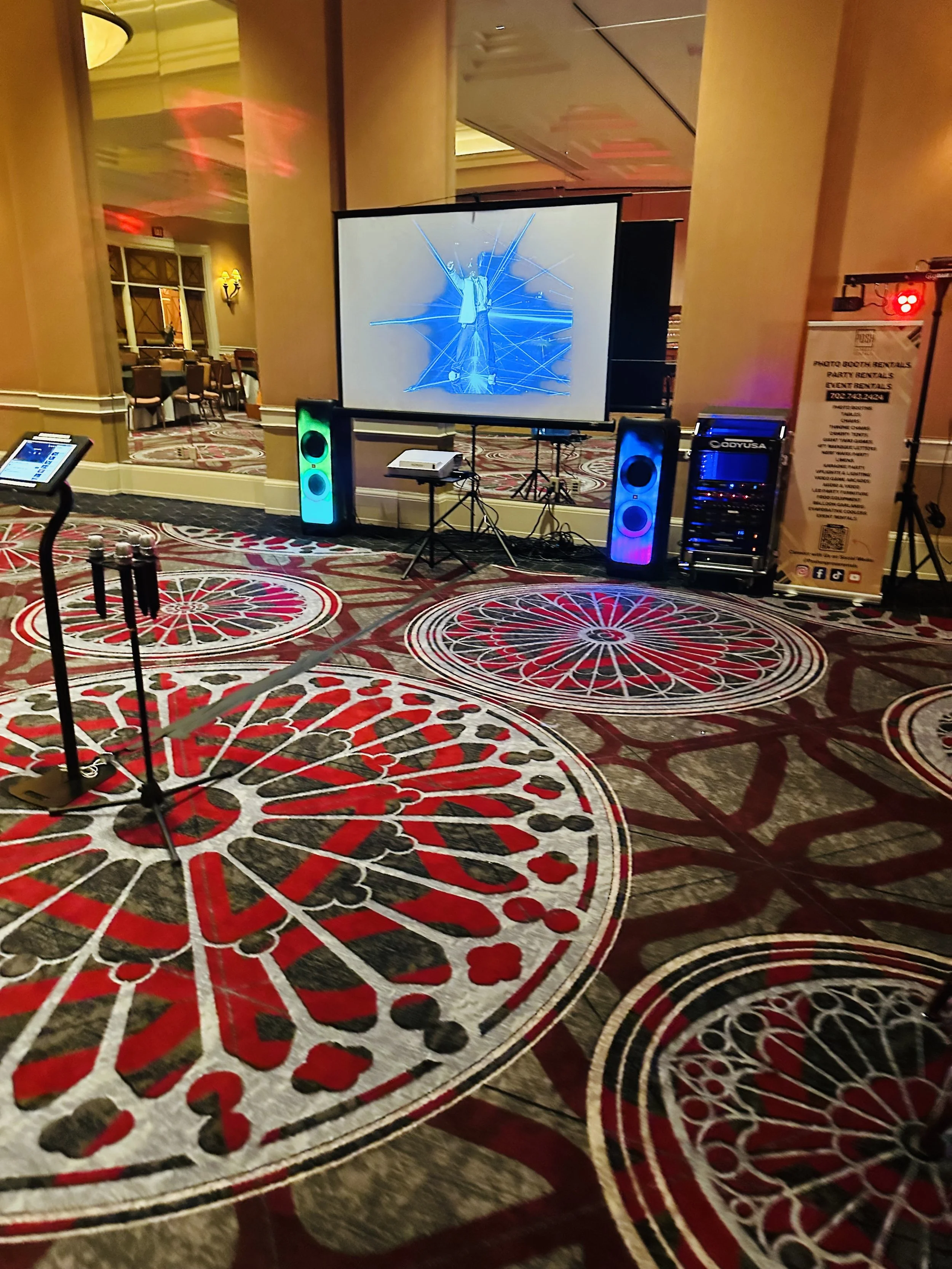 Indoor event space with a digital projector displaying graphics on a screen, colorful speakers, and ornate carpet design.