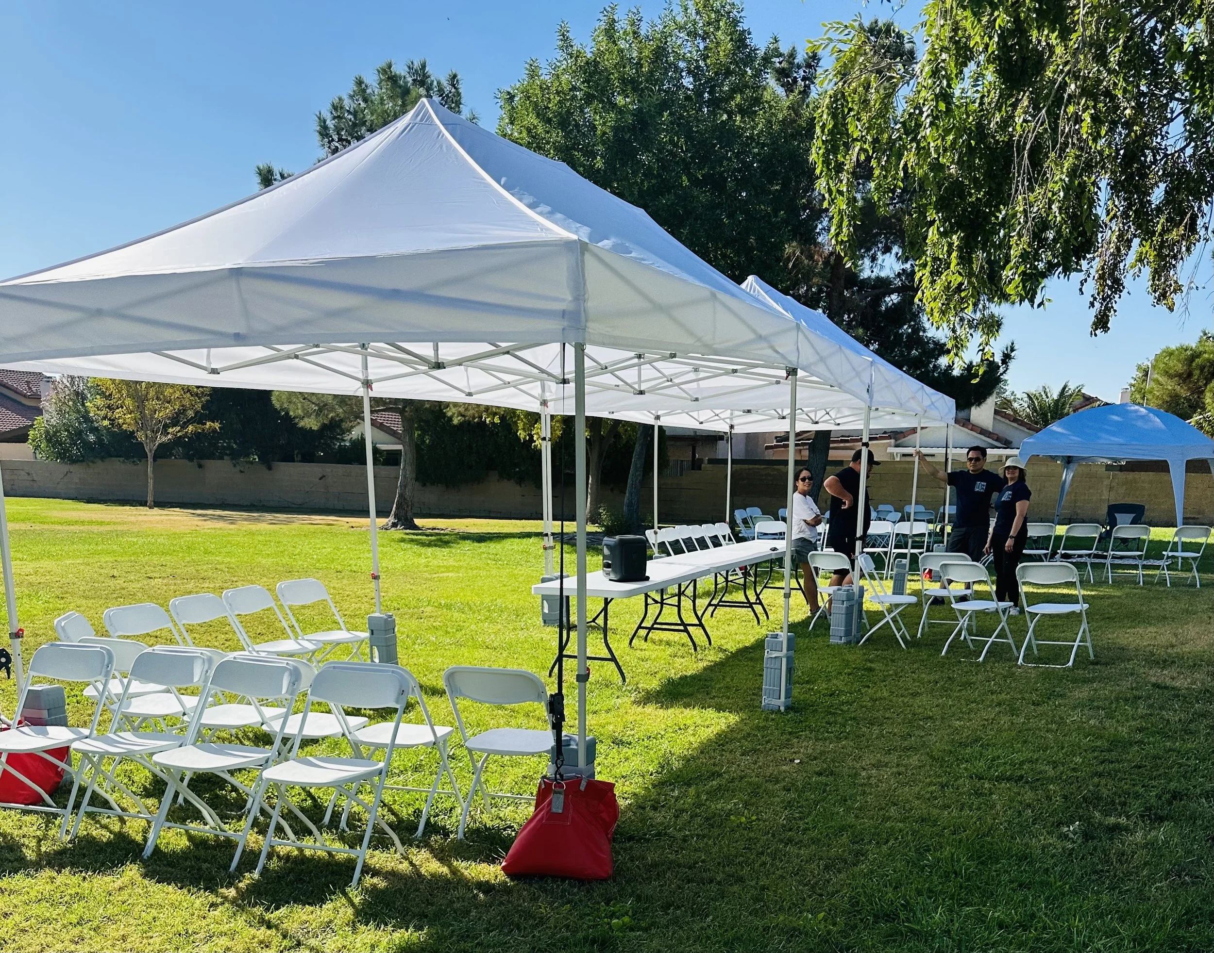 Outdoor event setup with white tents, folding chairs, and tables on a grassy area.