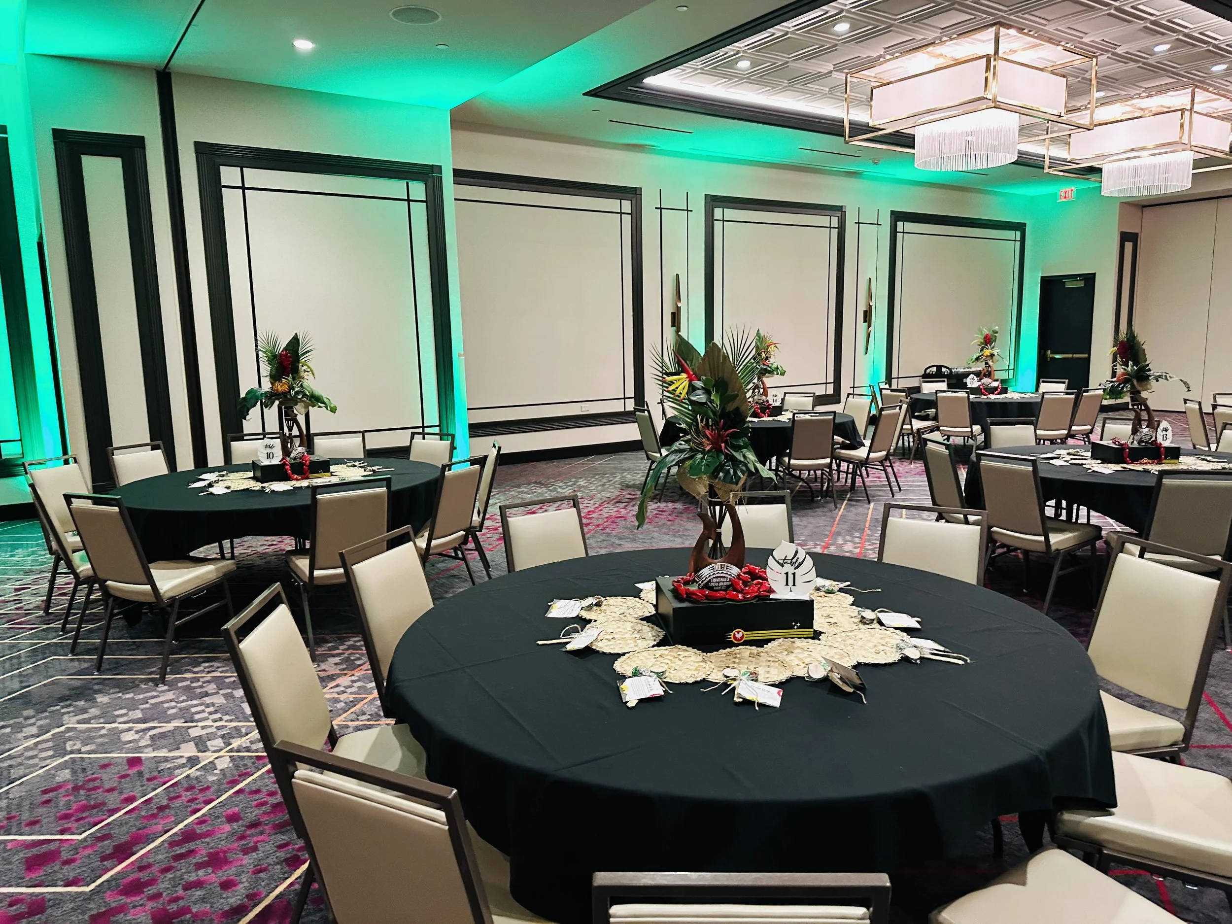 Decorated event room with round tables, black tablecloths, chairs, floral centerpieces, and green accent lighting.