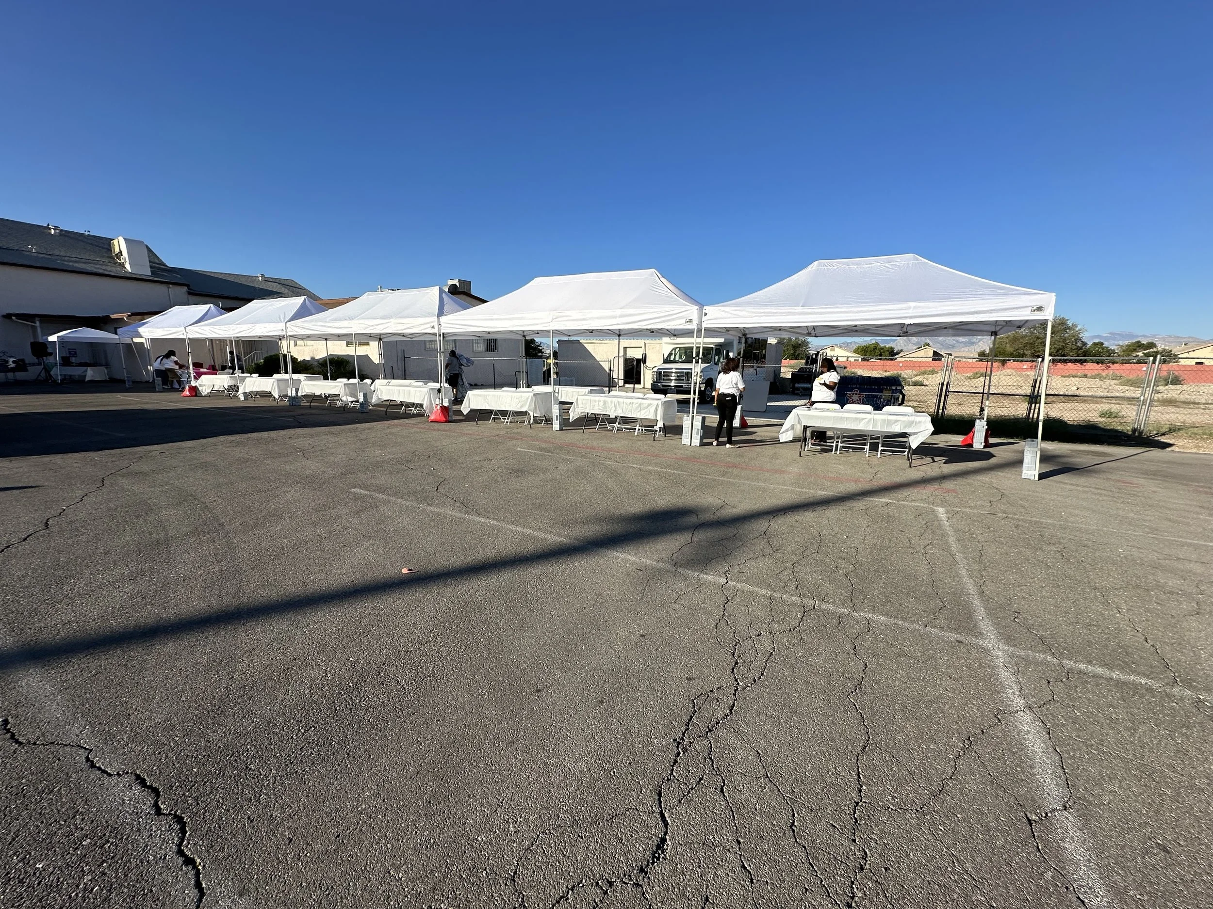 Outdoor event setup with white canopy tents, tables, and chairs in a paved area, clear blue sky, people setting up.