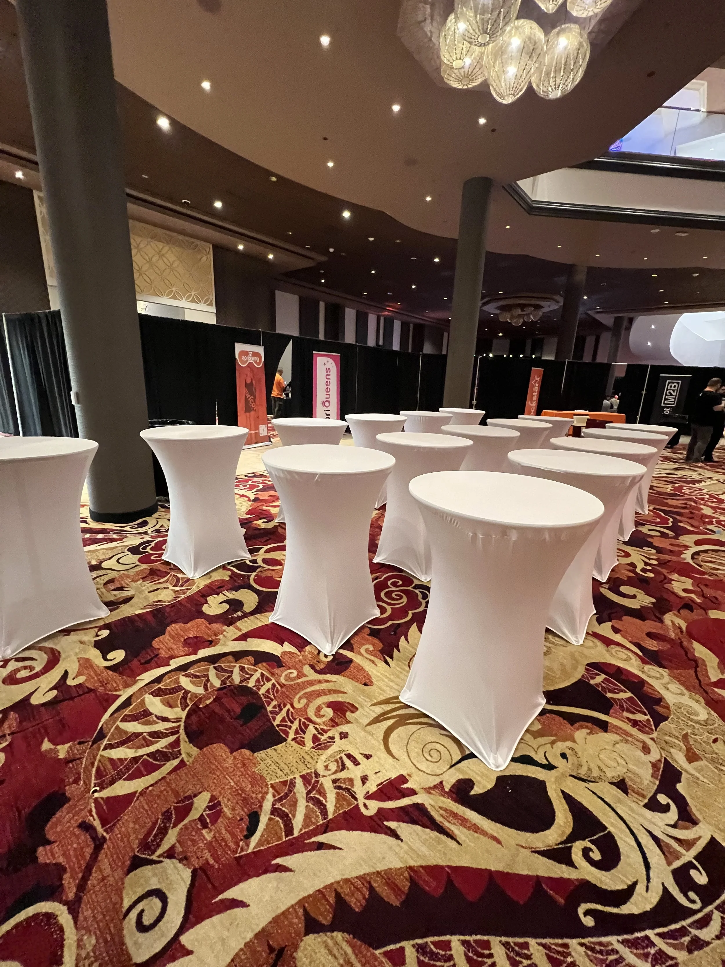 Hotel event space with high tables covered in white cloths, ornate carpet, and black drapes in background.