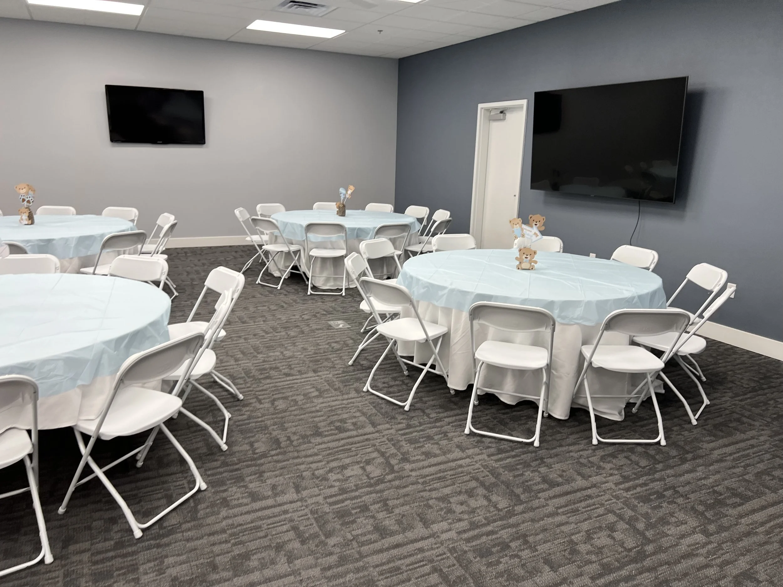 Conference room with round tables covered in light blue tablecloths, white folding chairs, teddy bear decorations, gray carpet, and two wall-mounted TVs.