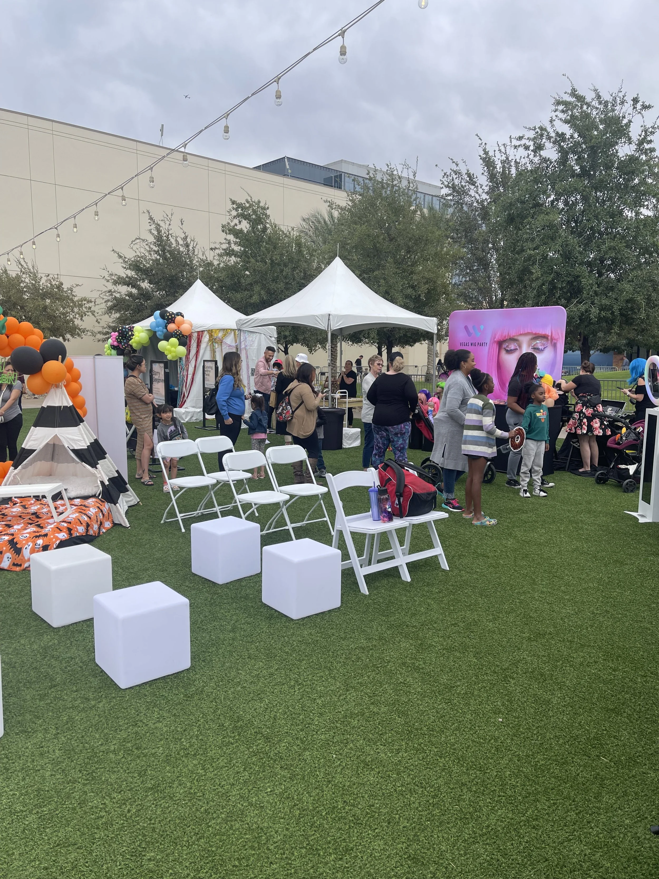 Outdoor event with white tents and people gathered, decorated with colorful balloons and string lights. Folding chairs and cube seating on grassy area.