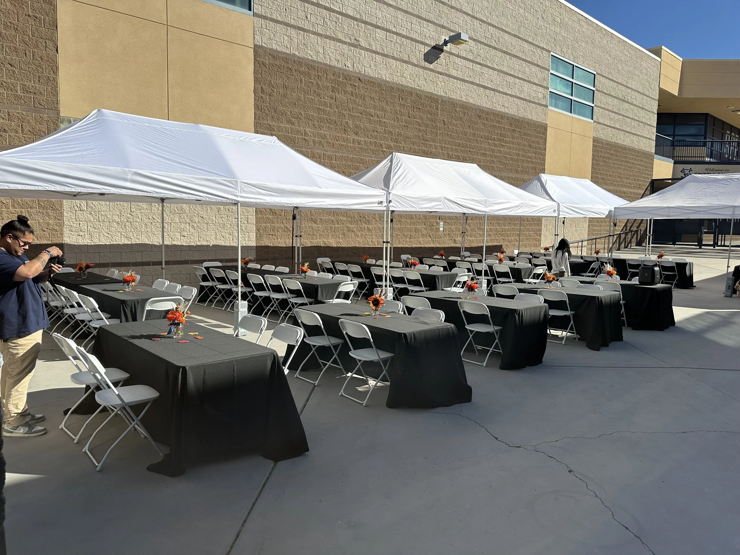 Outdoor event setup with white tents, tables covered in black tablecloths, white chairs, and floral centerpieces. Person taking photos.