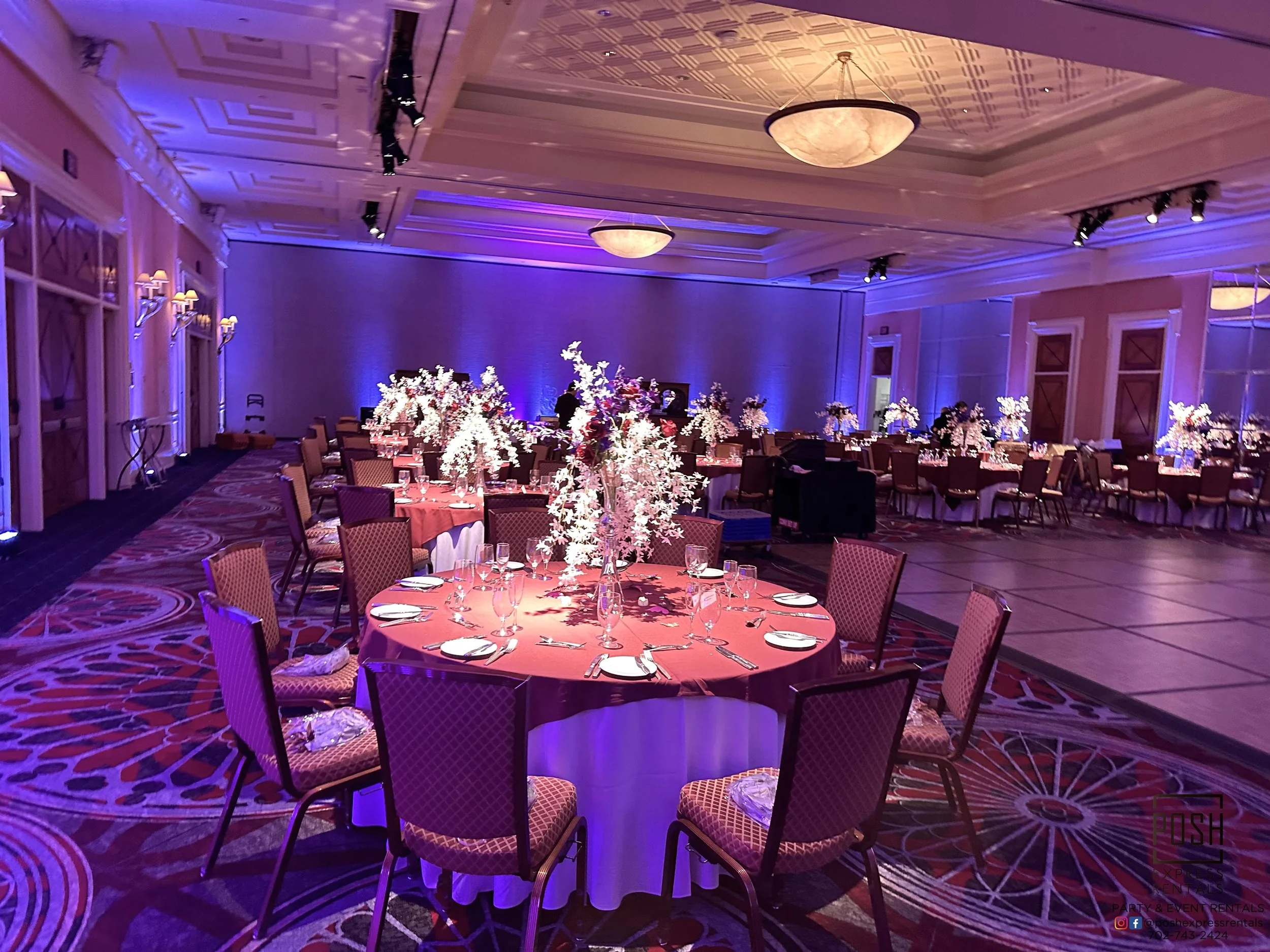 Lavishly decorated banquet hall with round tables and floral centerpieces, set for a formal event, featuring elegant lighting and a carpeted floor.