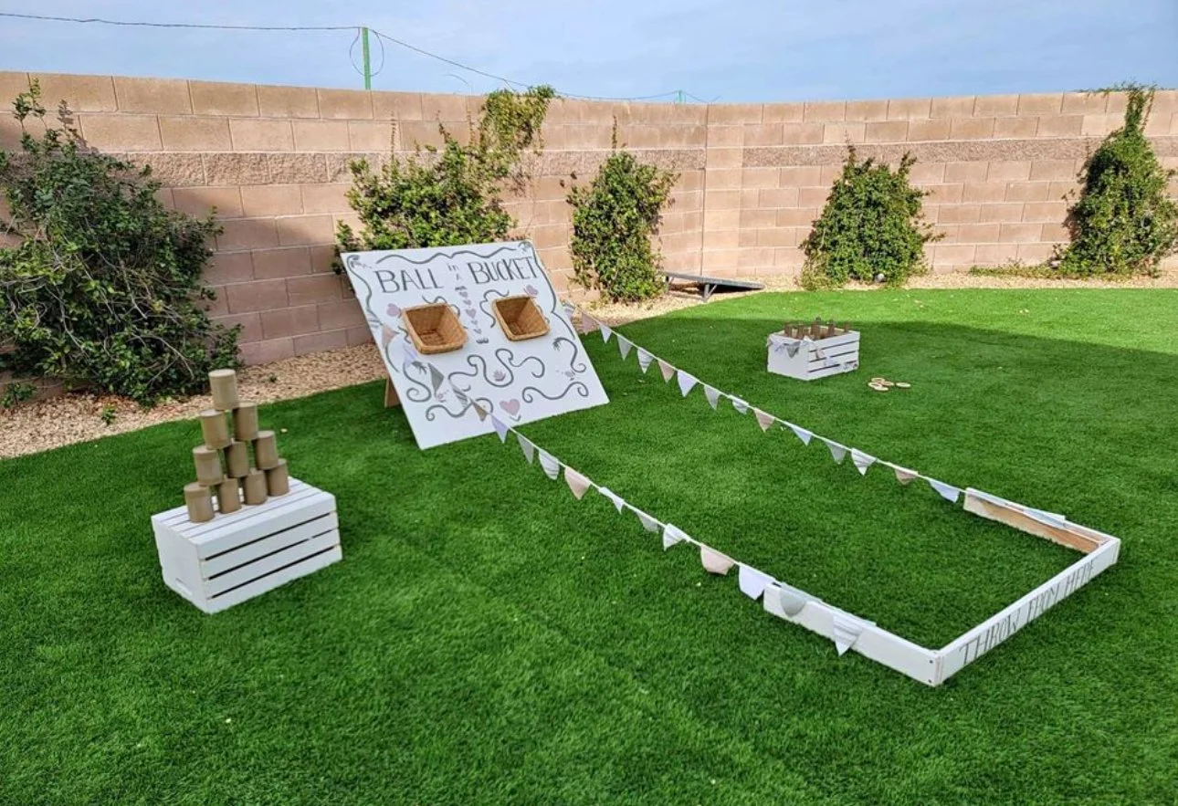 Outdoor carnival games setup on grass with a "Ball in Bucket" board, stacked cans, and fenced area.