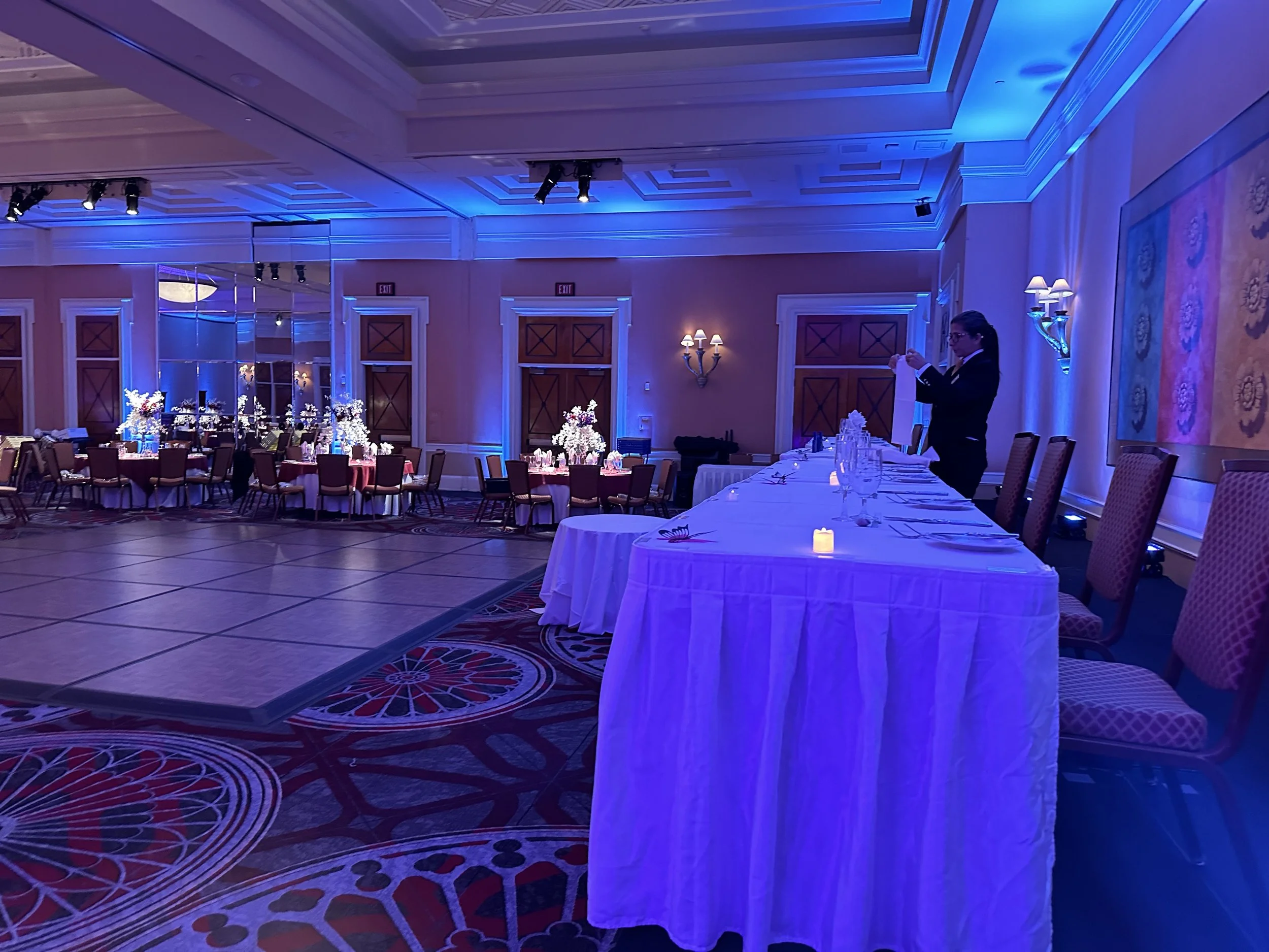 Elegant banquet hall setup with tables, chairs, and decorations, featuring blue lighting and a person arranging a table setting.
