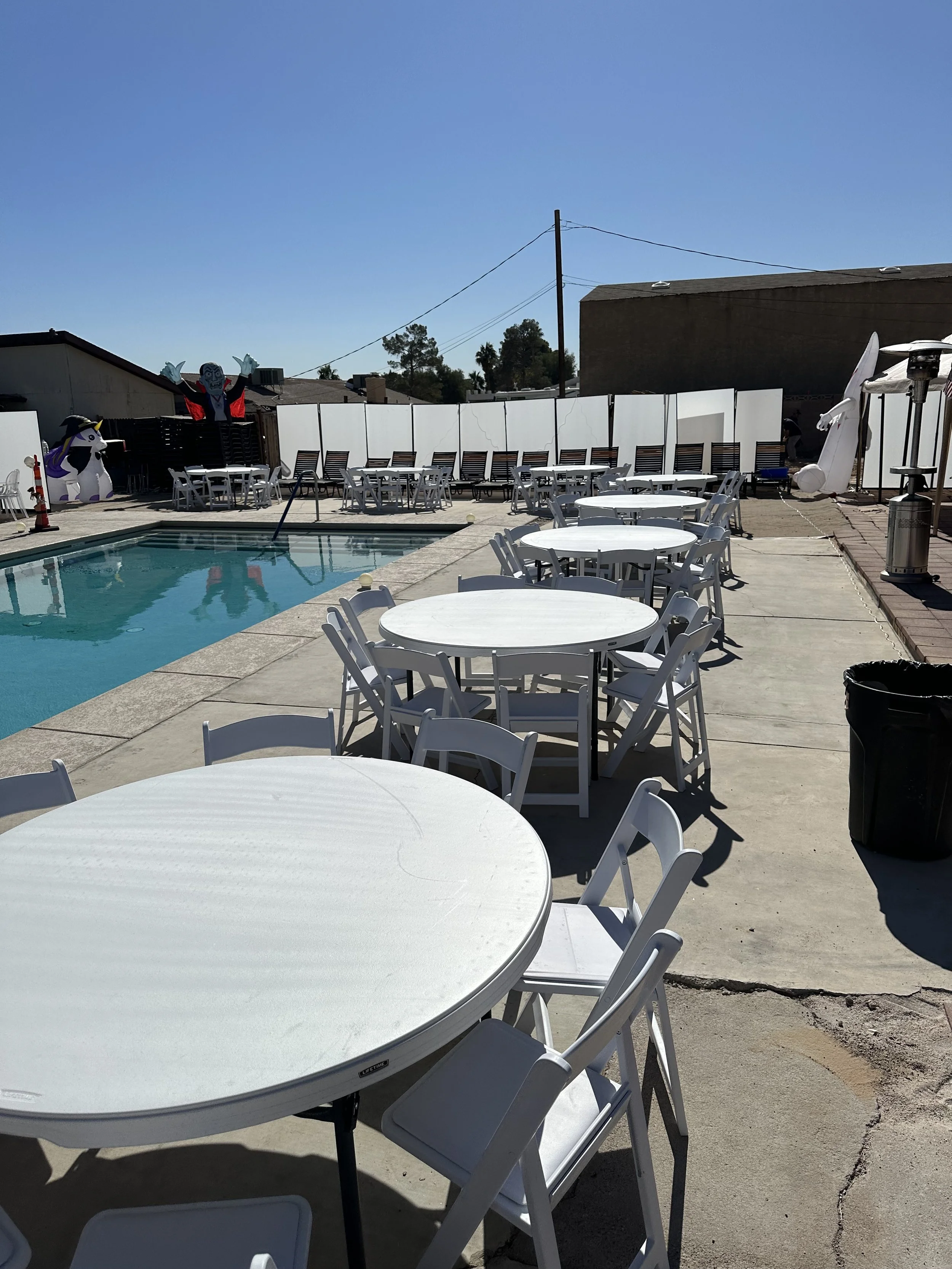 Poolside setup with round tables and white chairs, inflatable Halloween decorations, and outdoor umbrellas on a sunny day.