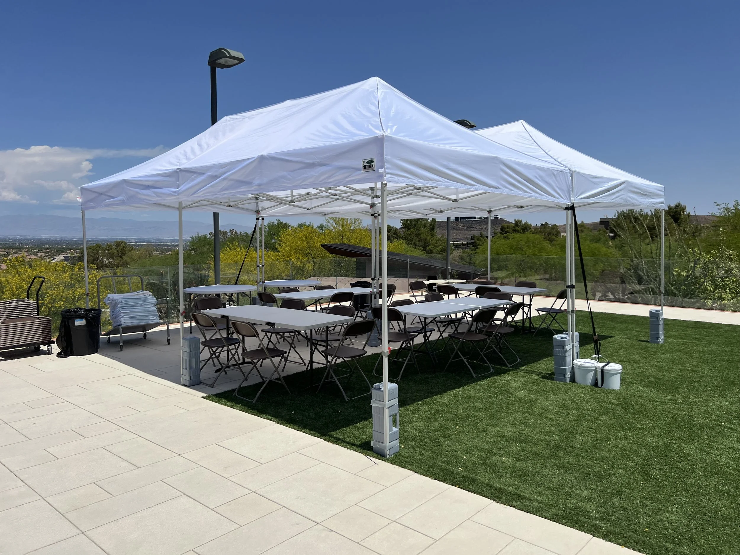 Outdoor setup with white canopy tents, folding tables, and chairs on grassy and tiled area, under clear sky.
