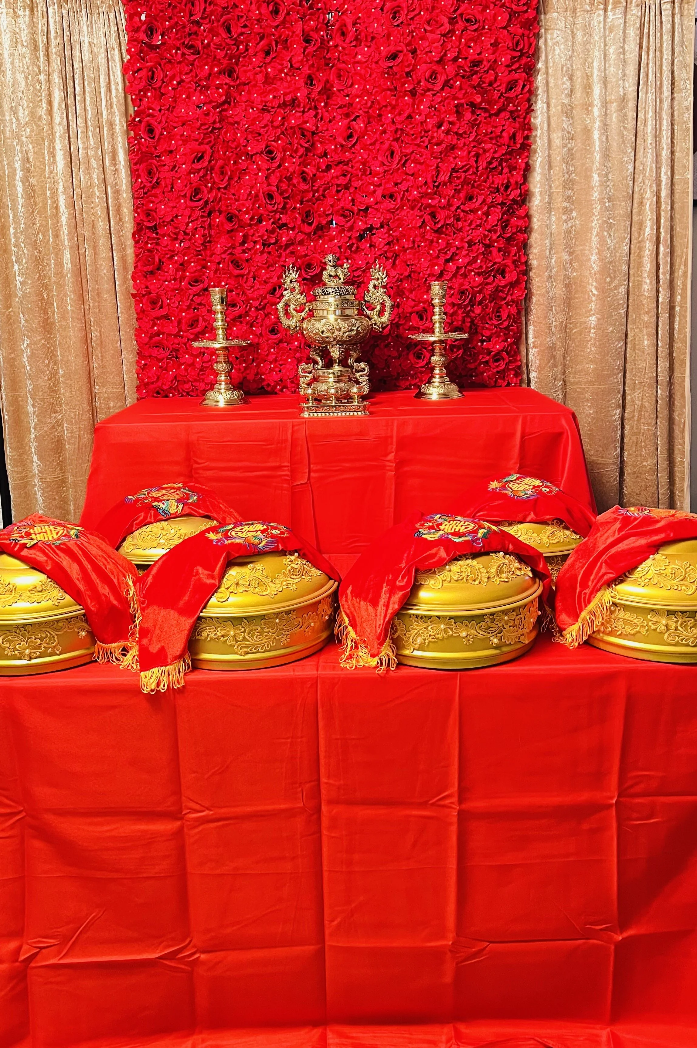 Traditional Asian ceremonial setup with gold and red decorations, ornate containers, cushions, and red floral backdrop.