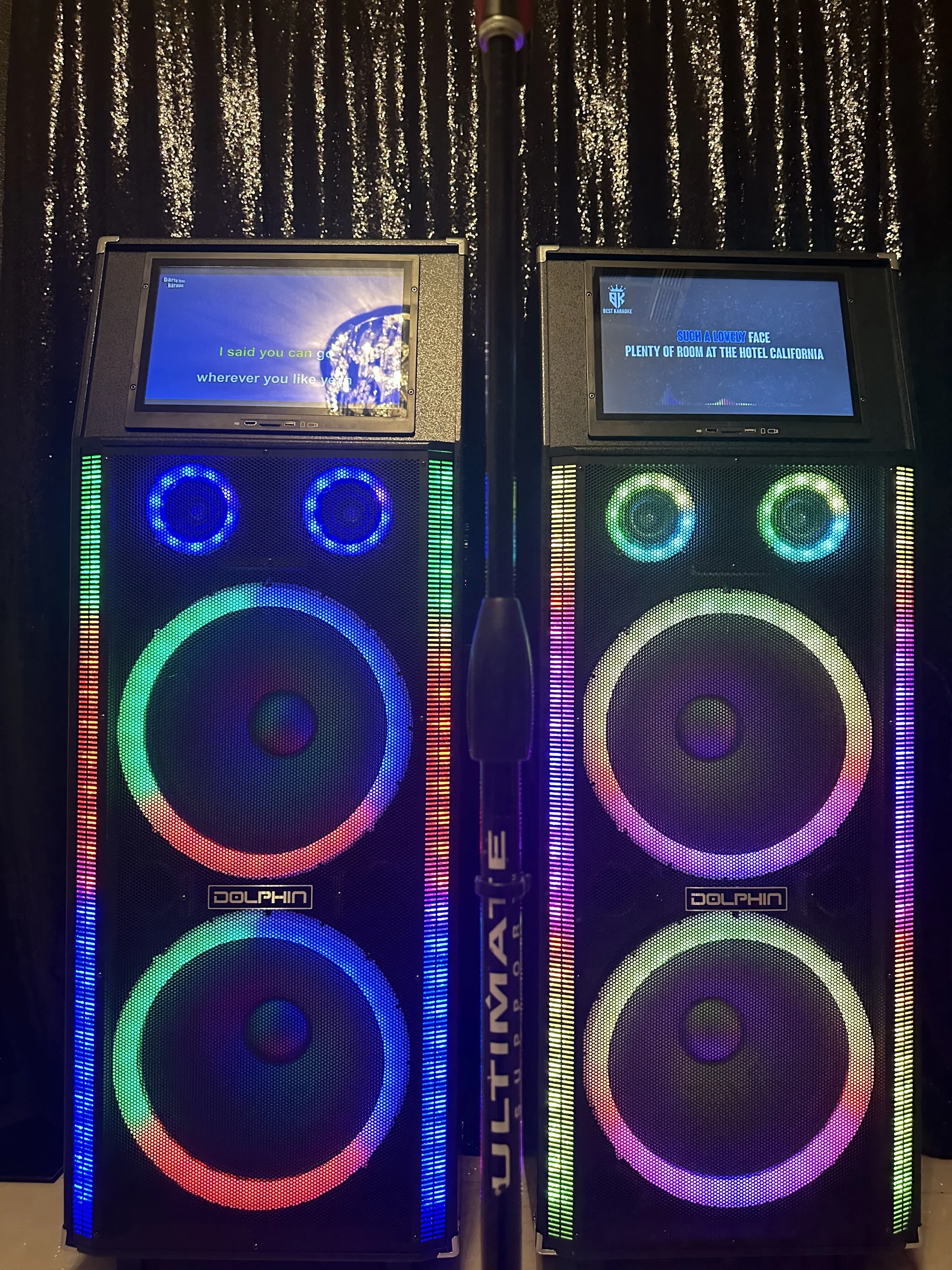 Two large speakers with colorful LED lights and screens displaying karaoke lyrics, separated by a microphone stand, set against a glittery black curtain.