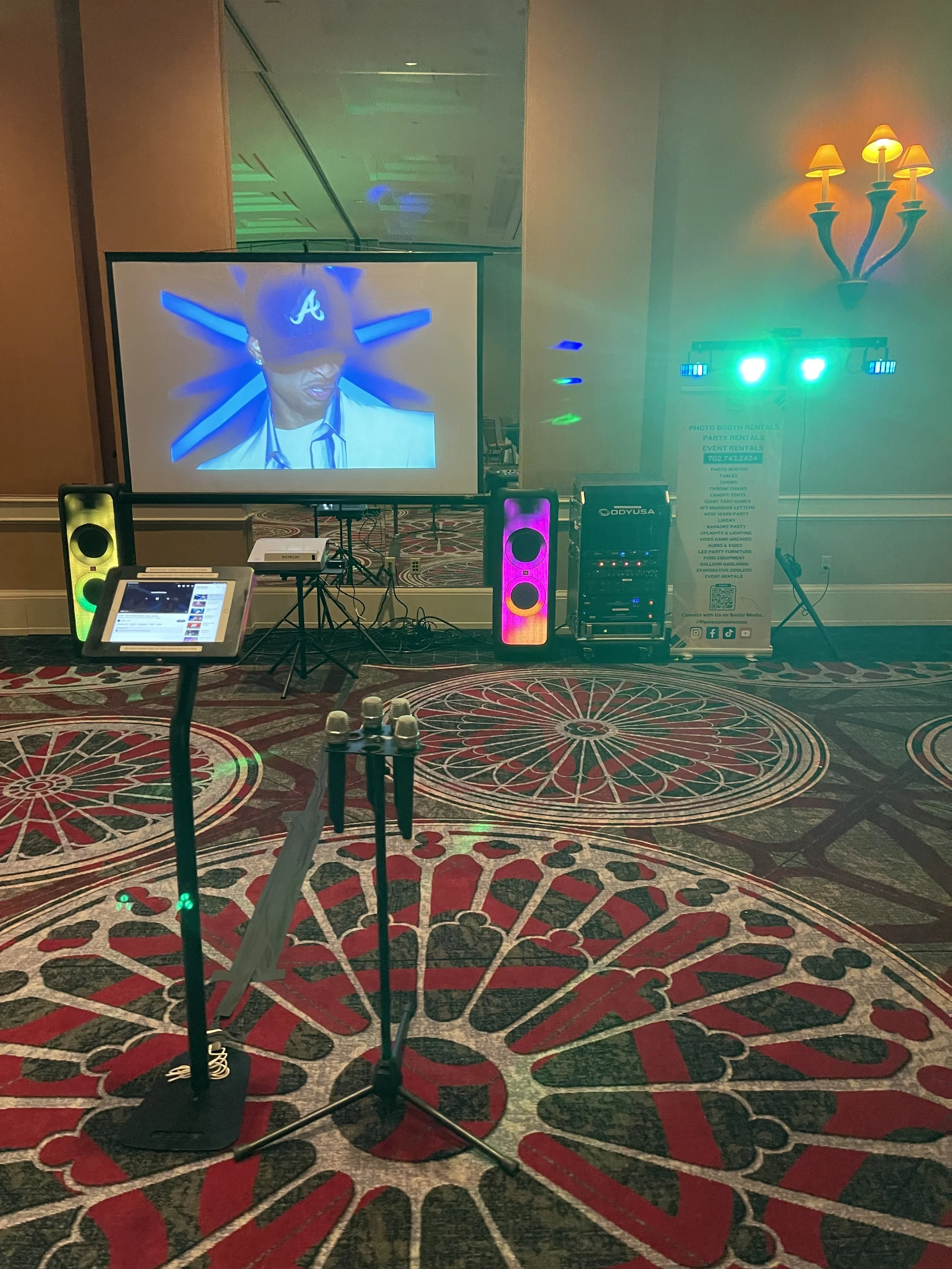 Event room with karaoke setup, featuring a screen displaying a person, speakers, microphone stand, LED lights, and DJ equipment on patterned carpet.