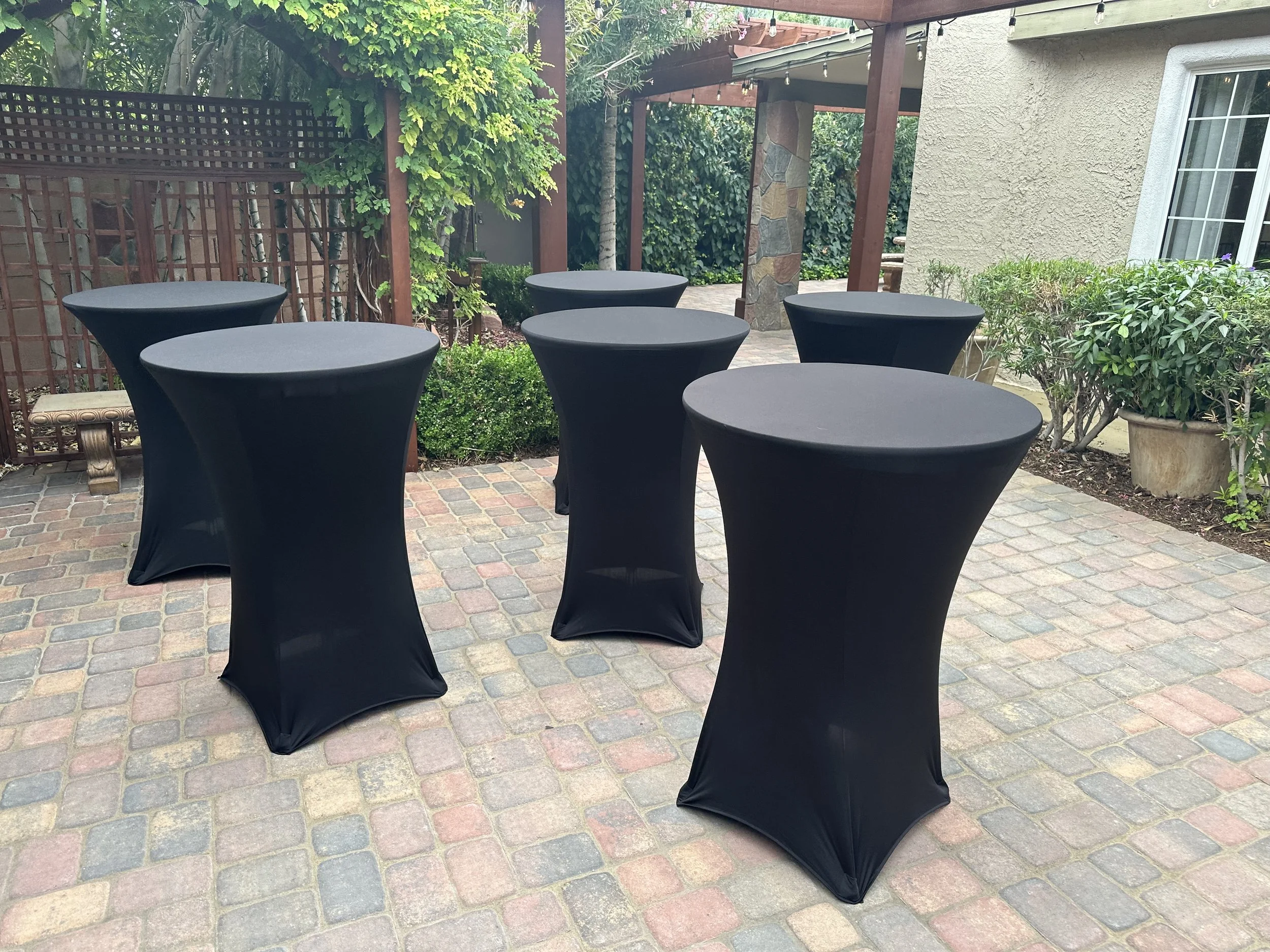 Outdoor patio with six black standing cocktail tables, brick flooring, greenery, and pergola in the background.