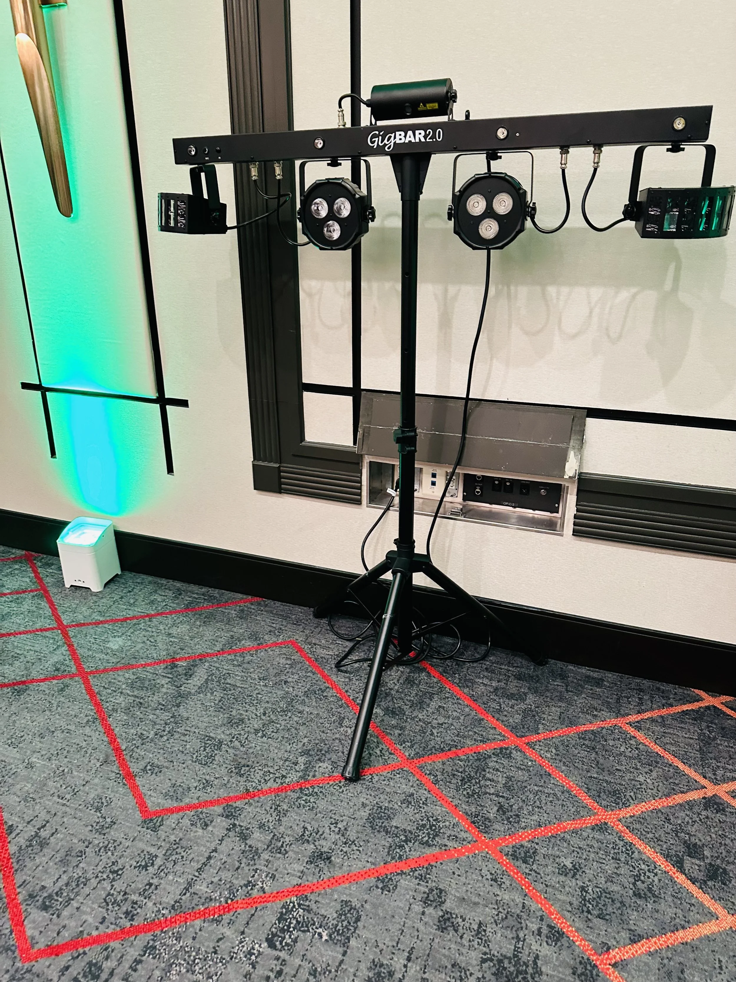 DJ lighting setup featuring GigBAR 2.0 with tripod stand in a room with patterned carpet and green accent lights.