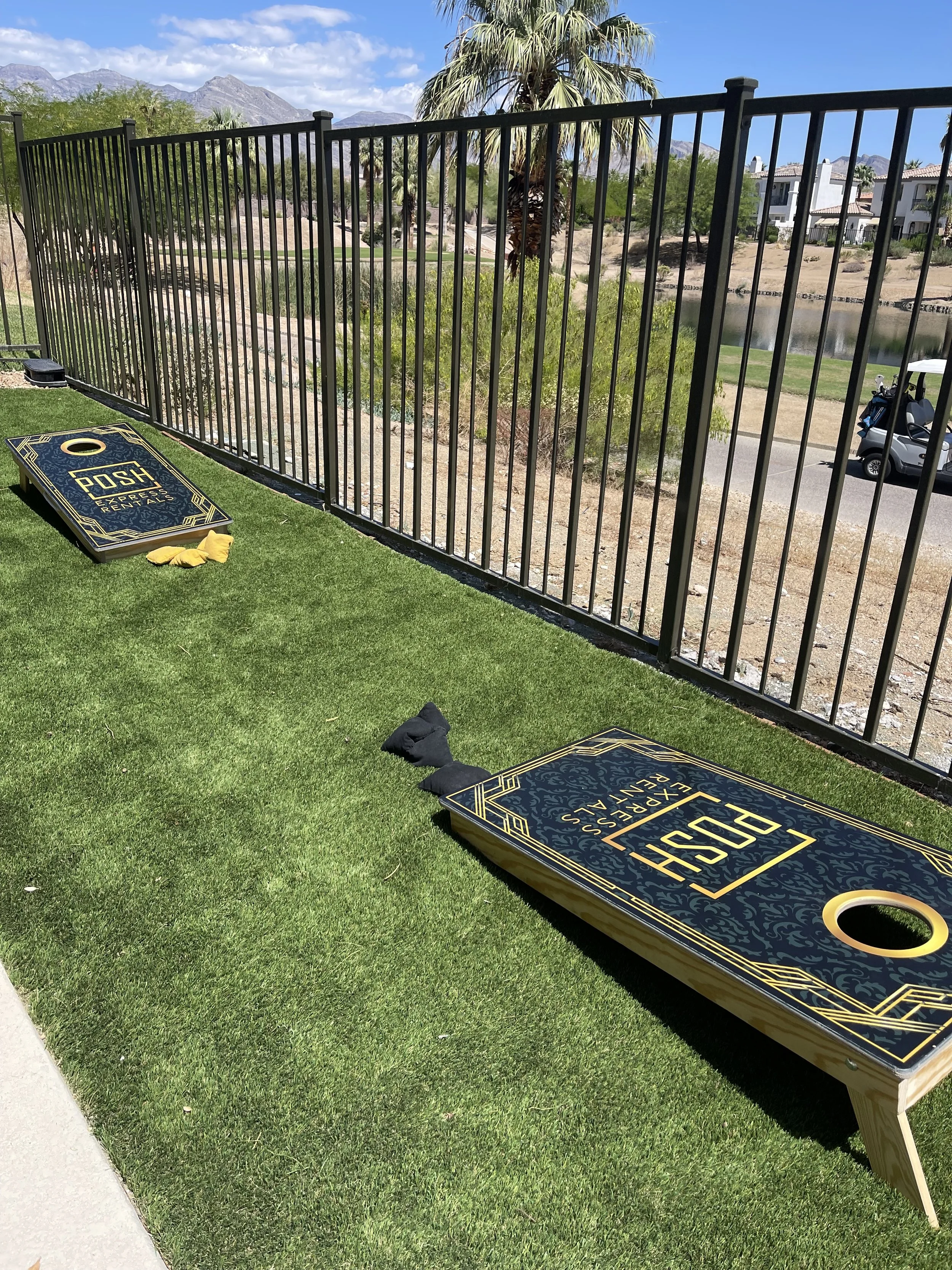 Cornhole boards on artificial grass with a metal fence and palm trees in the background.
