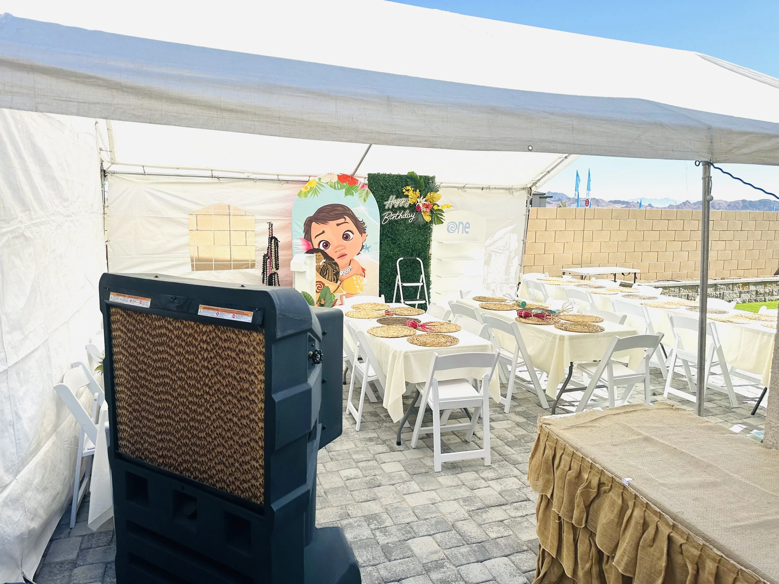 Outdoor birthday party setup with tent, decorated tables, backdrop with cartoon character, and patio cooling fan.
