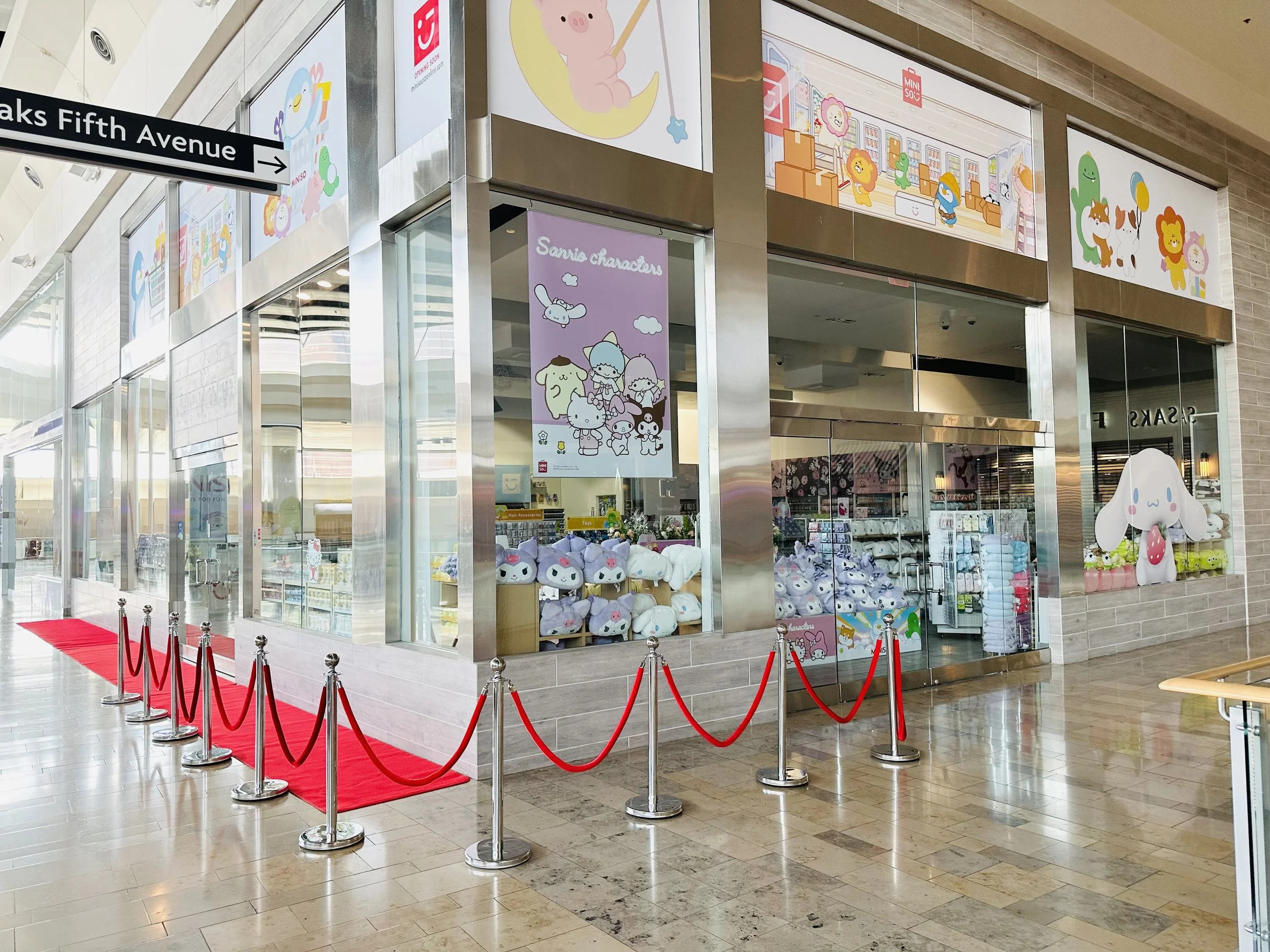 Storefront with cartoon characters, plush toys on display, red carpet ropes, and signs indicating location near Saks Fifth Avenue.