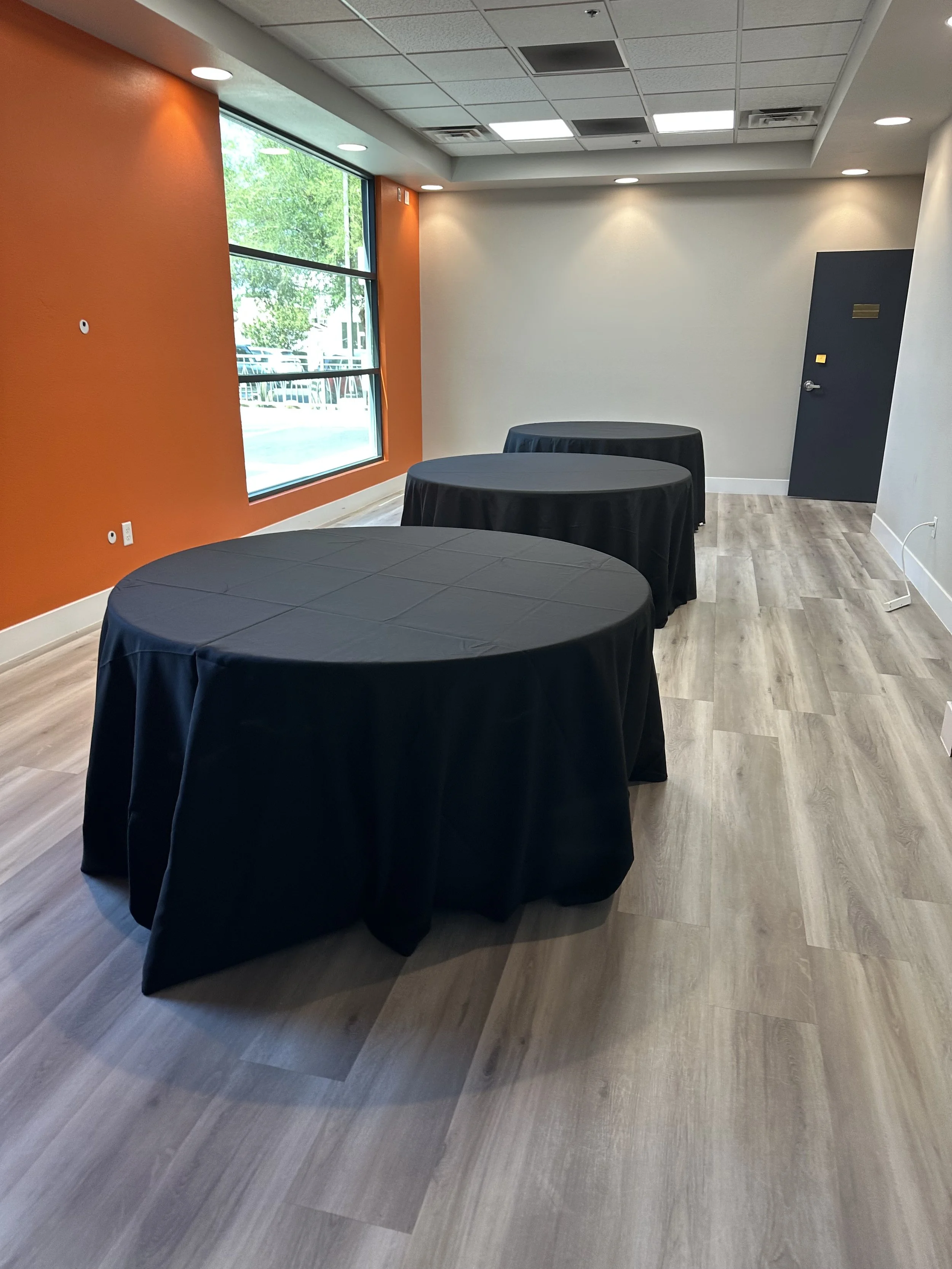Empty room with round tables covered in black tablecloths, orange accent wall, large window, and wood flooring.