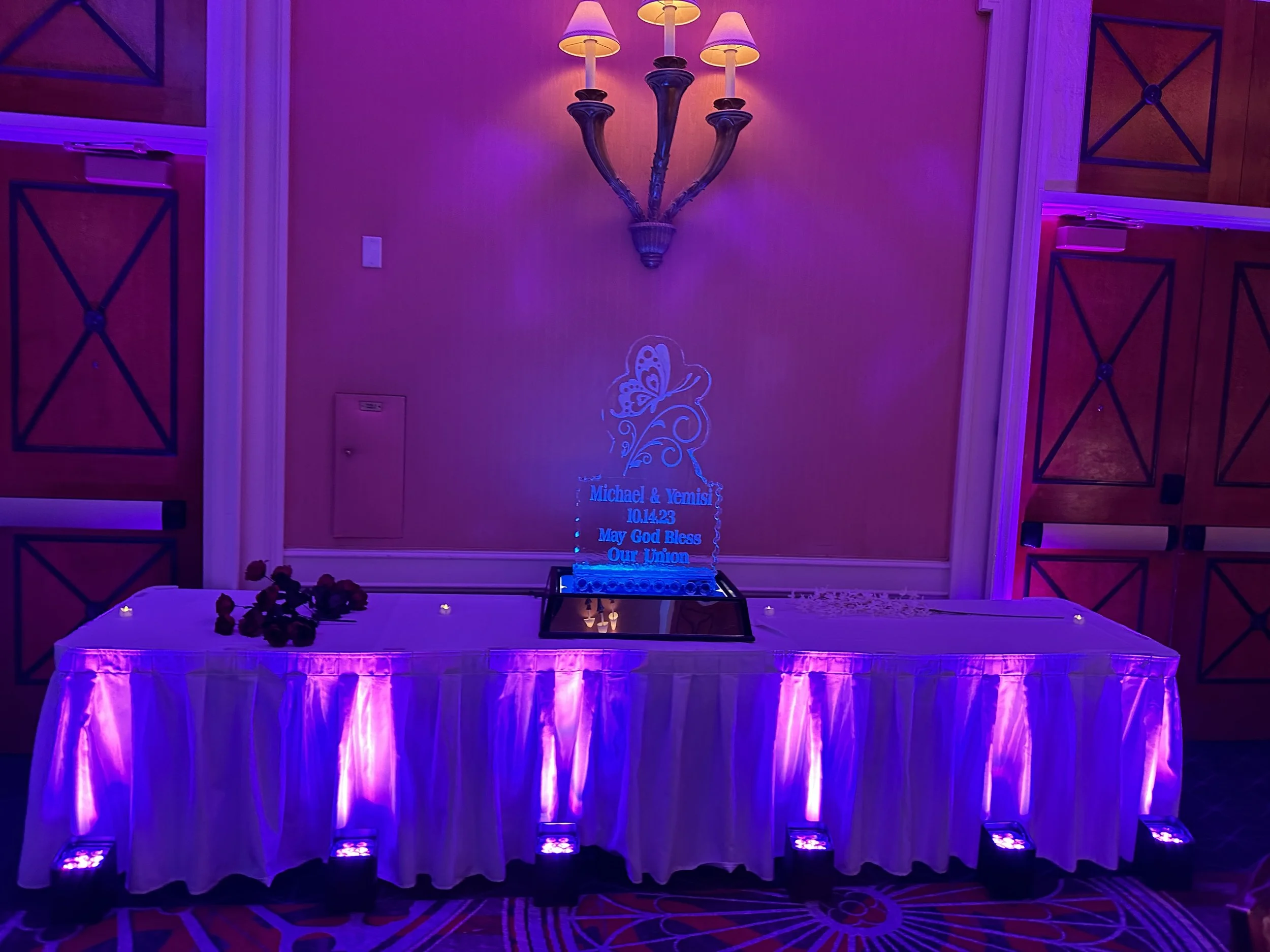 Decorative wedding table display with illuminated sign, roses, and purple lighting.