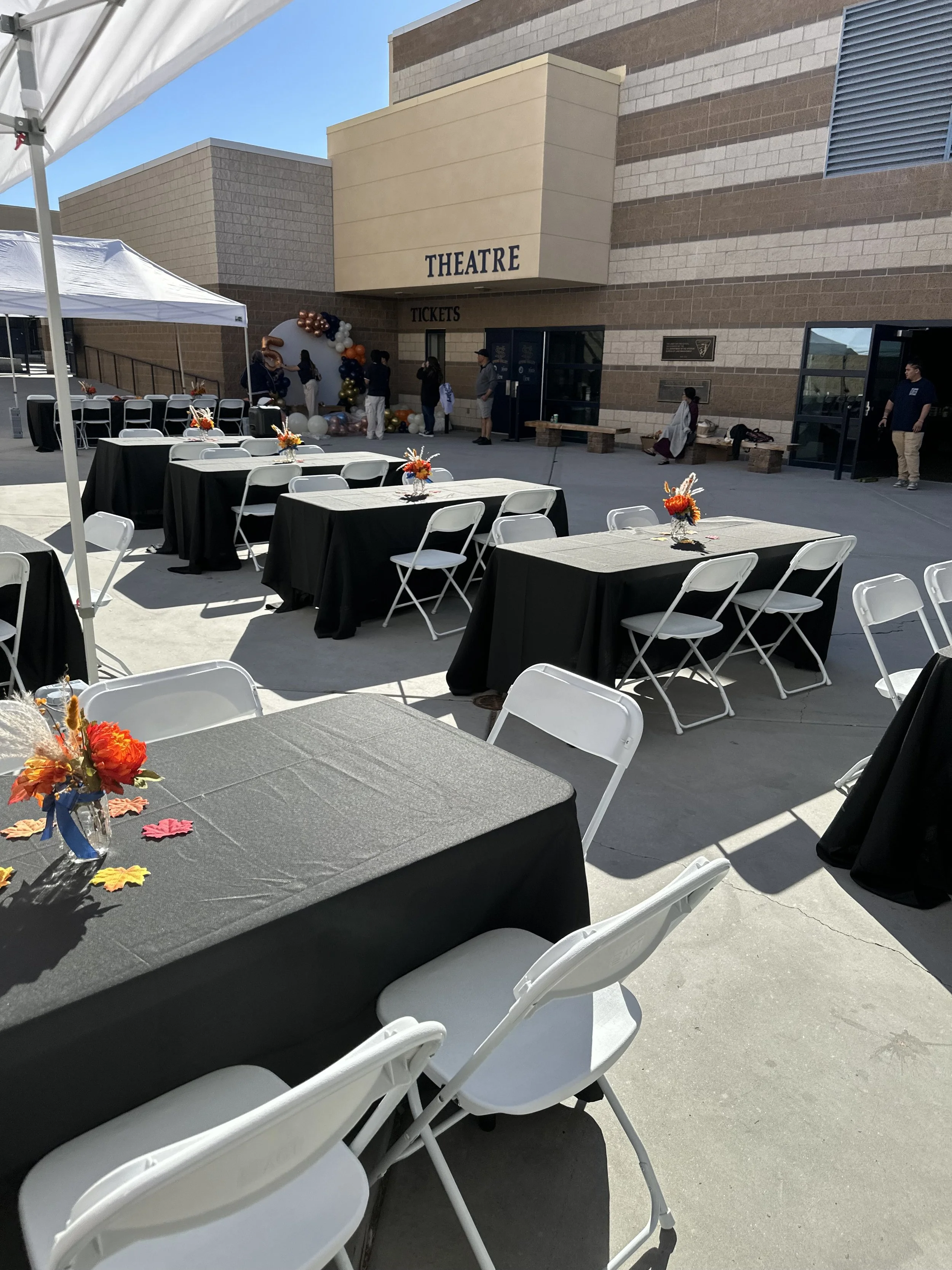 Outdoor event space with tables and chairs decorated with black tablecloths and floral centerpieces, near a theater entrance.