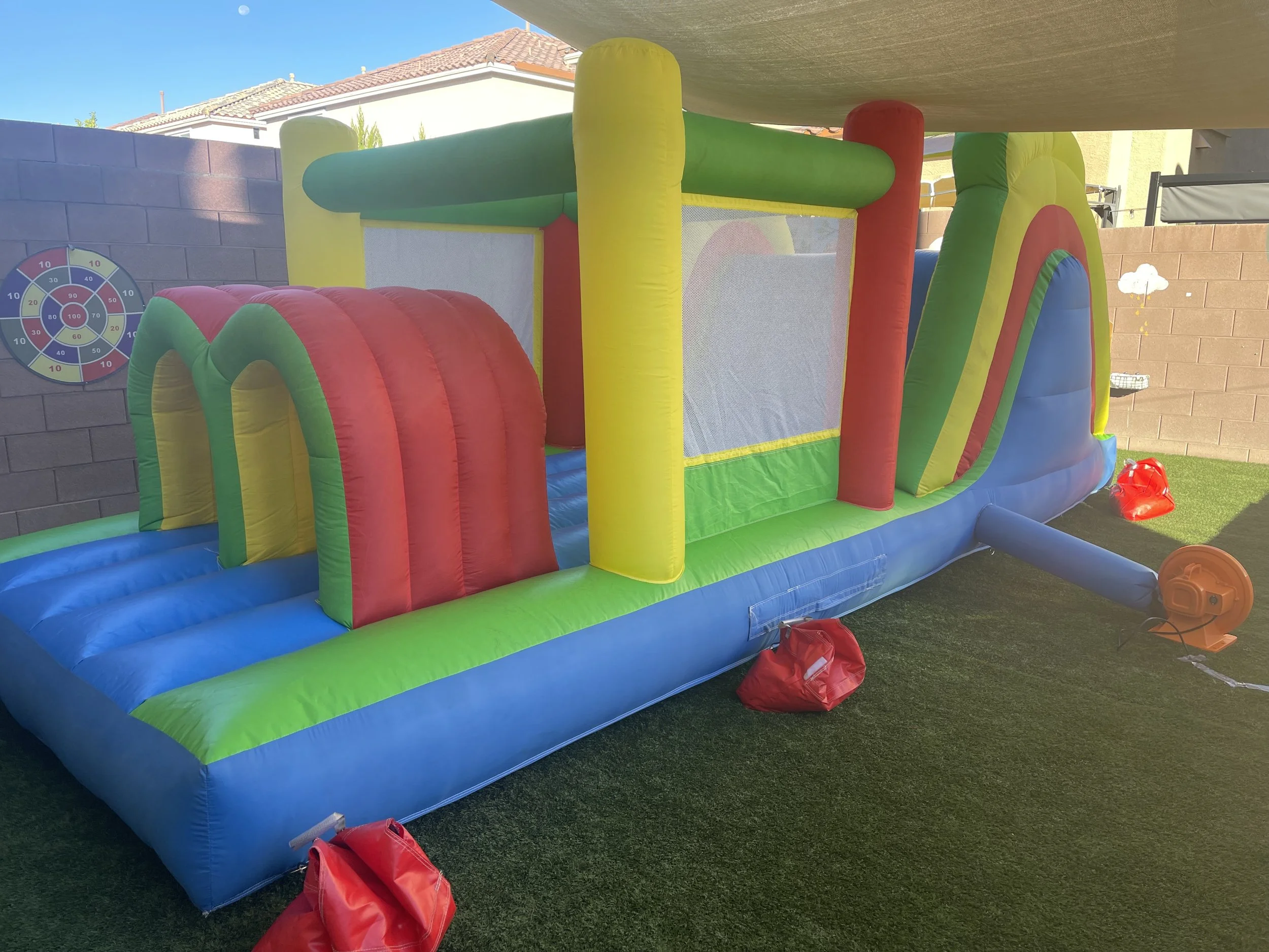 Colorful inflatable bounce house on artificial grass in a backyard, with a safety net and deflated red sandbags for stability.