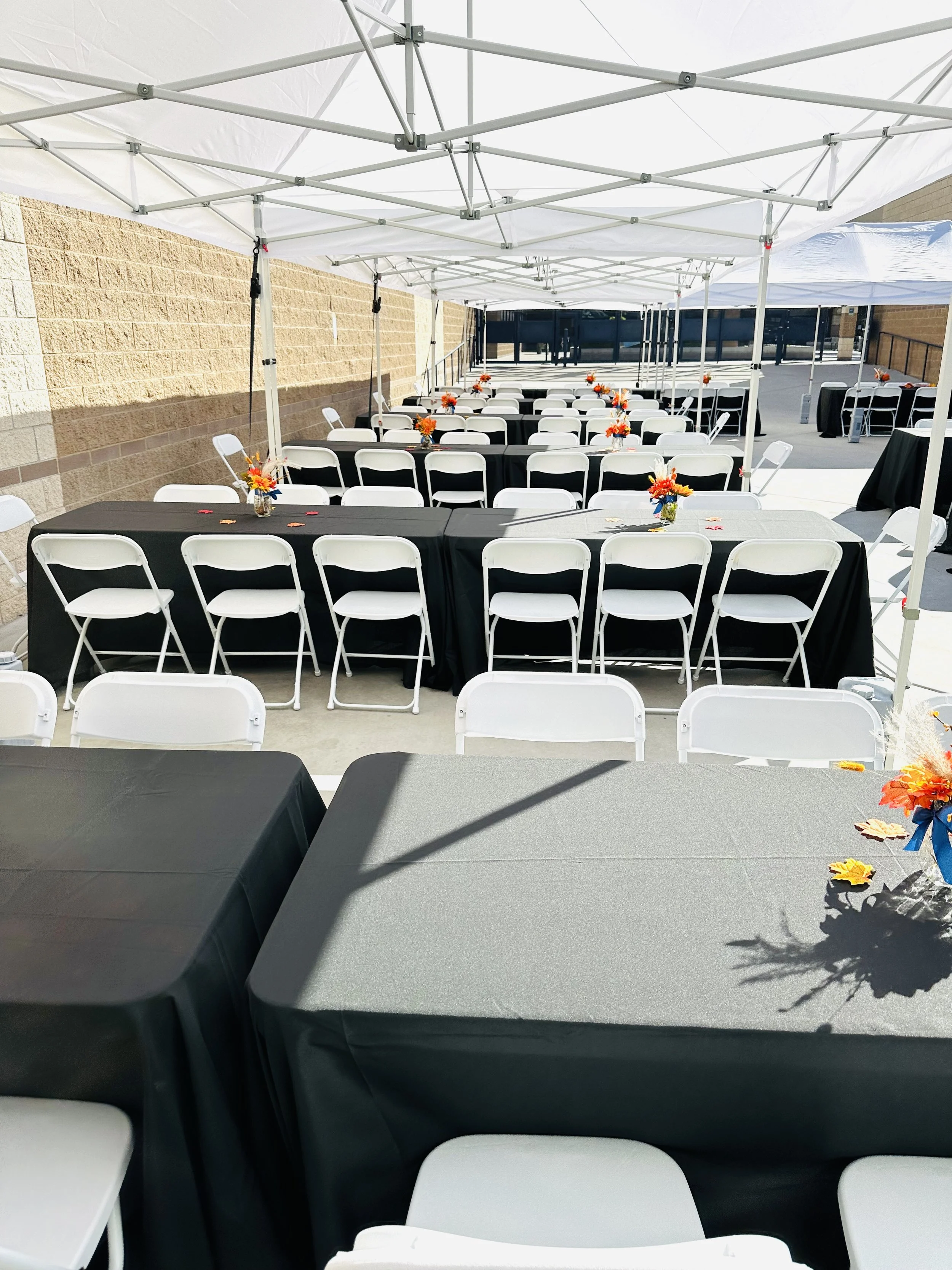 Outdoor event setup with black tablecloths, white chairs, and fall decorations under canopy tents.