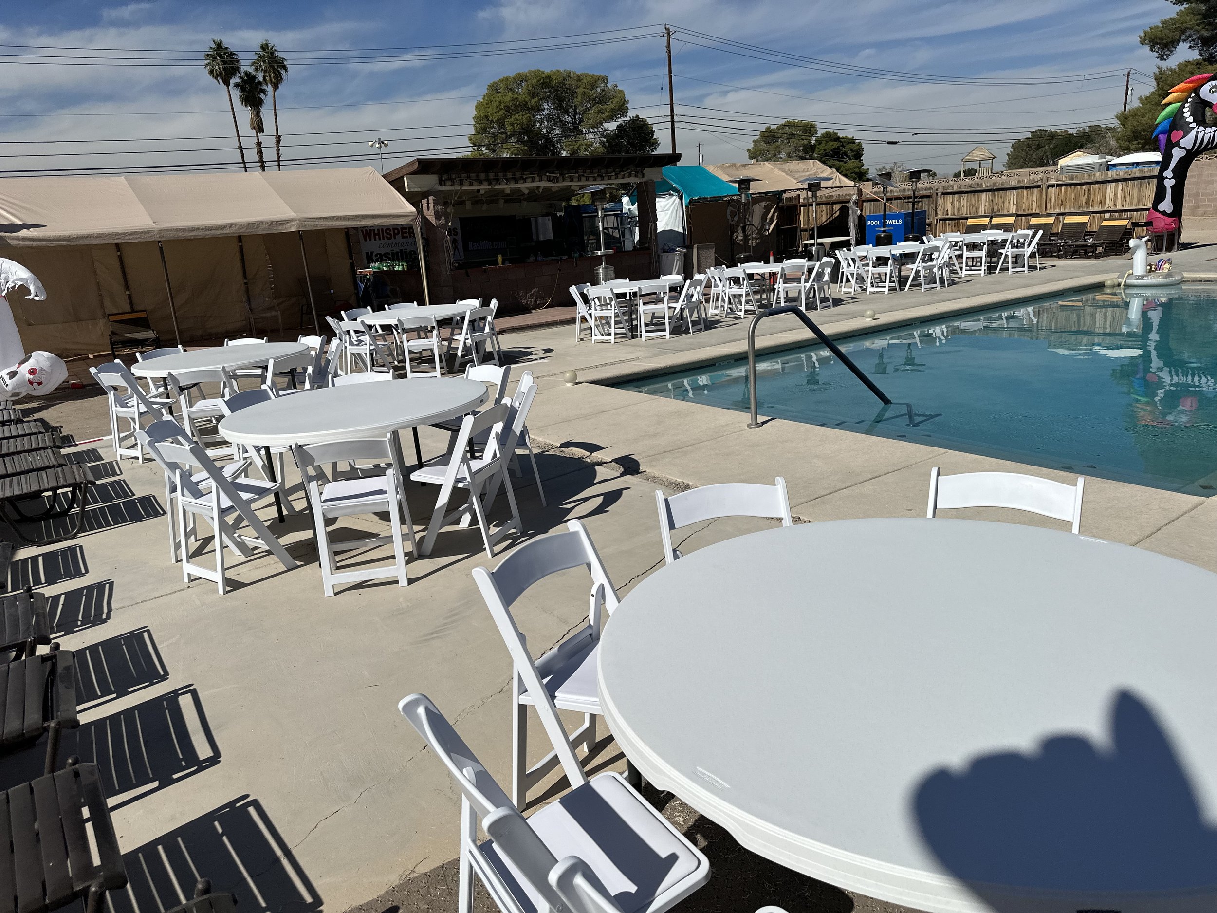 Outdoor pool area with white round tables and chairs, surrounded by tents and wooden structures, under a sunny sky with palm trees.