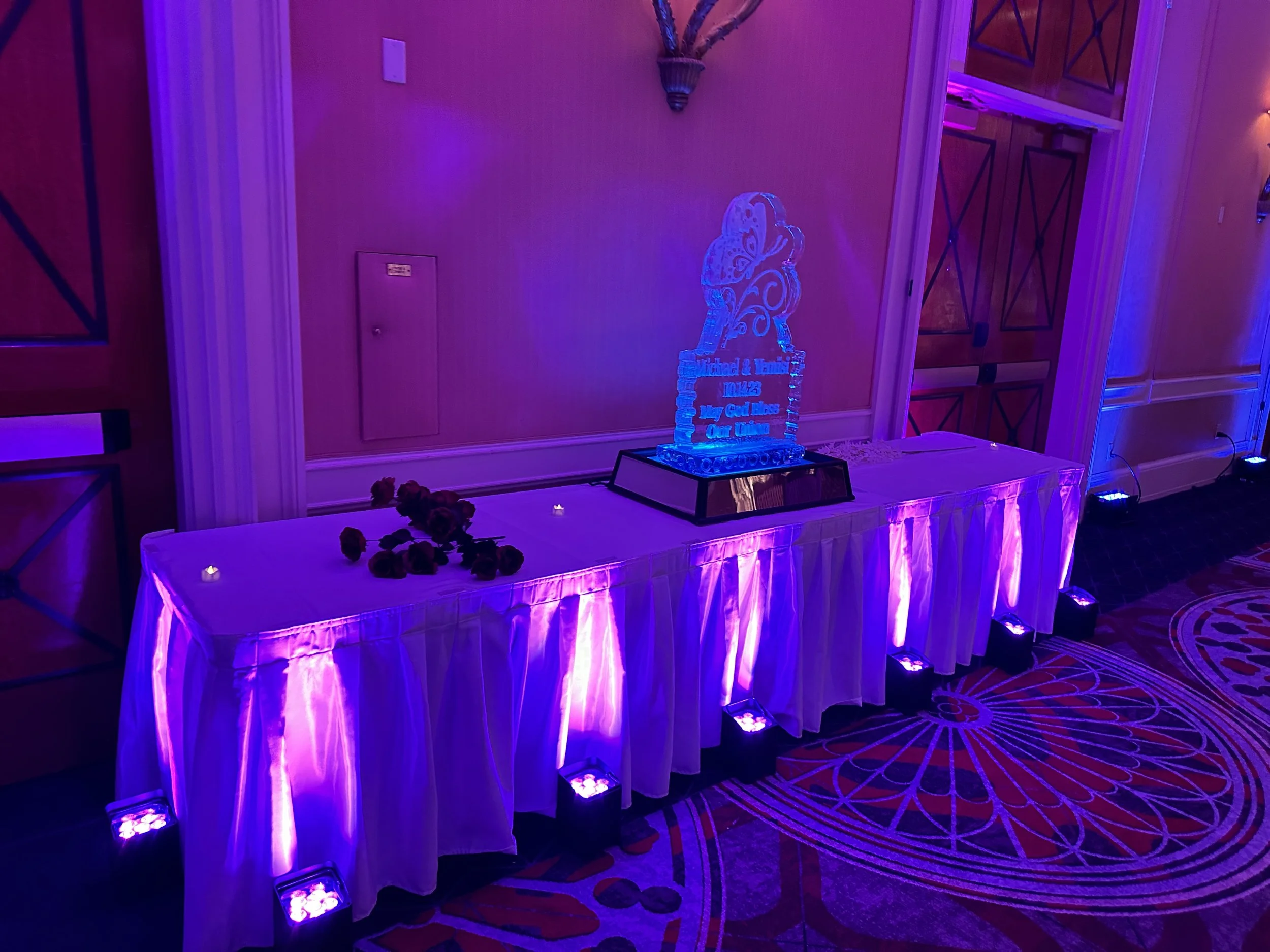Wedding reception table setup with purple lighting, featuring an ice sculpture, roses on the table, and decorative candles.