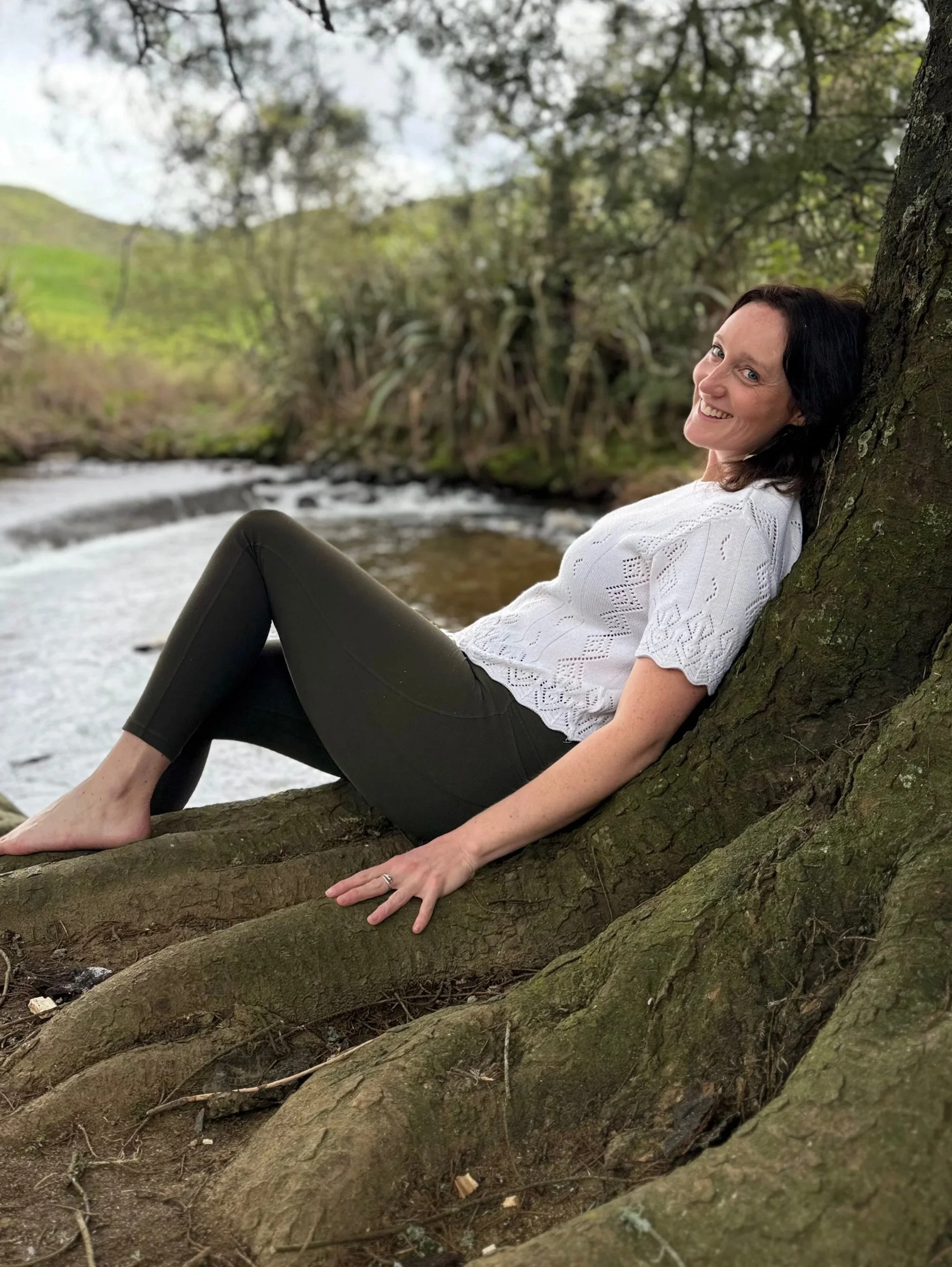 A woman with black hair, wearing a white blouse and black pants, relaxing on a tree root near a river with green hills and trees in the background.