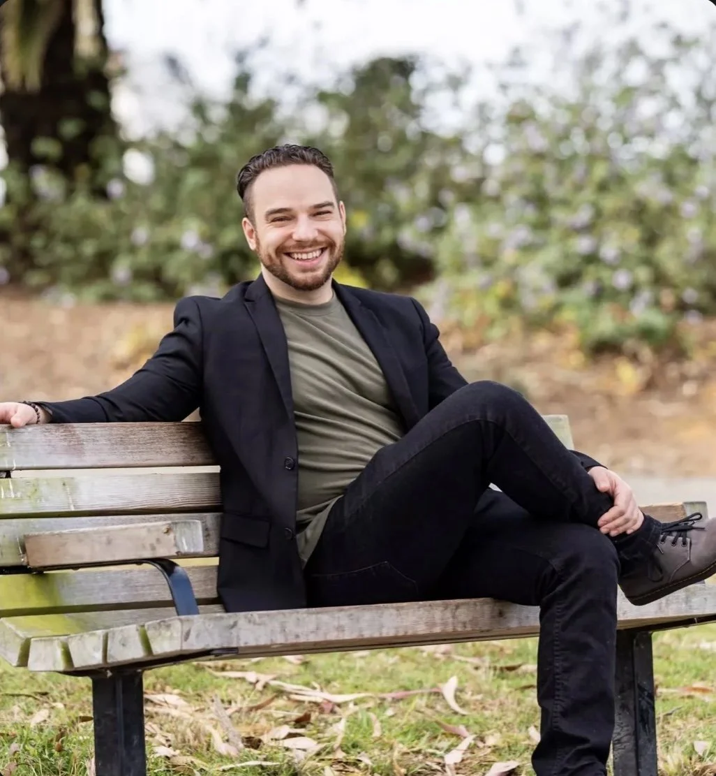 A smiling man with a beard sitting on a park bench outdoors, wearing a dark blazer and black pants, with greenery in the background.