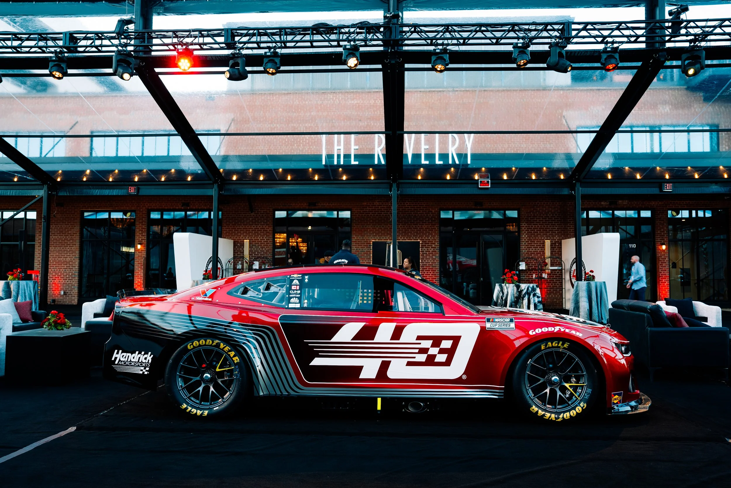 A red NASCAR race car with the number 43 and sponsors displayed, showcased indoors with seating areas, plants, and a sign reading 'THE RIVERY' in the background.