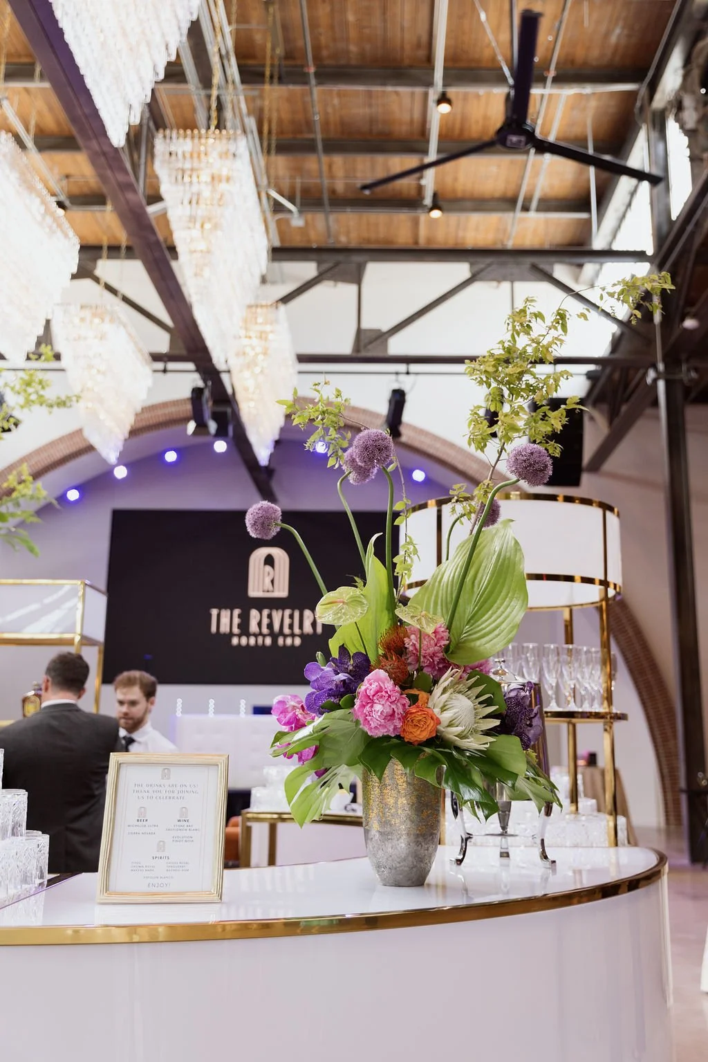 Elegant floral centerpiece with purple, pink, orange, and white flowers on a bar counter at an upscale event with chandeliers, a ceiling fan, and a black sign that reads 'The Revelry Sound Co' in the background.