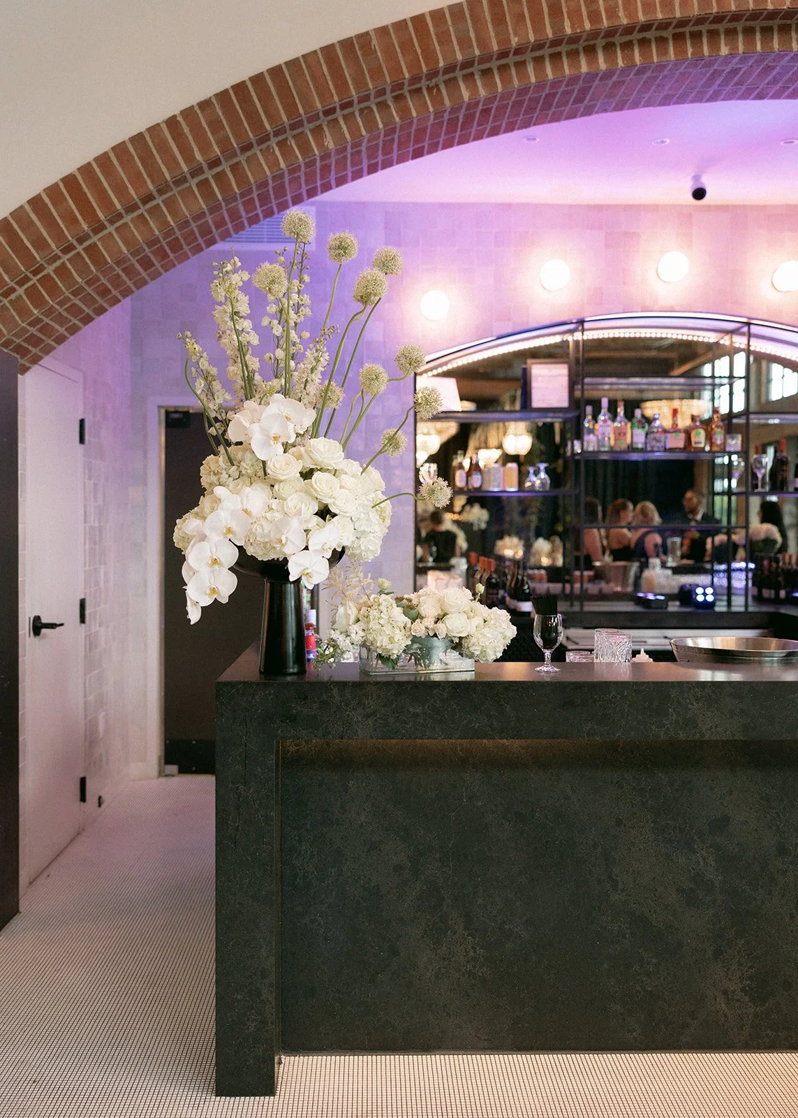 A black marble bar counter with a tall bouquet of white flowers in a black vase, set in an elegant restaurant or bar with exposed brick arches and warm lighting behind.
