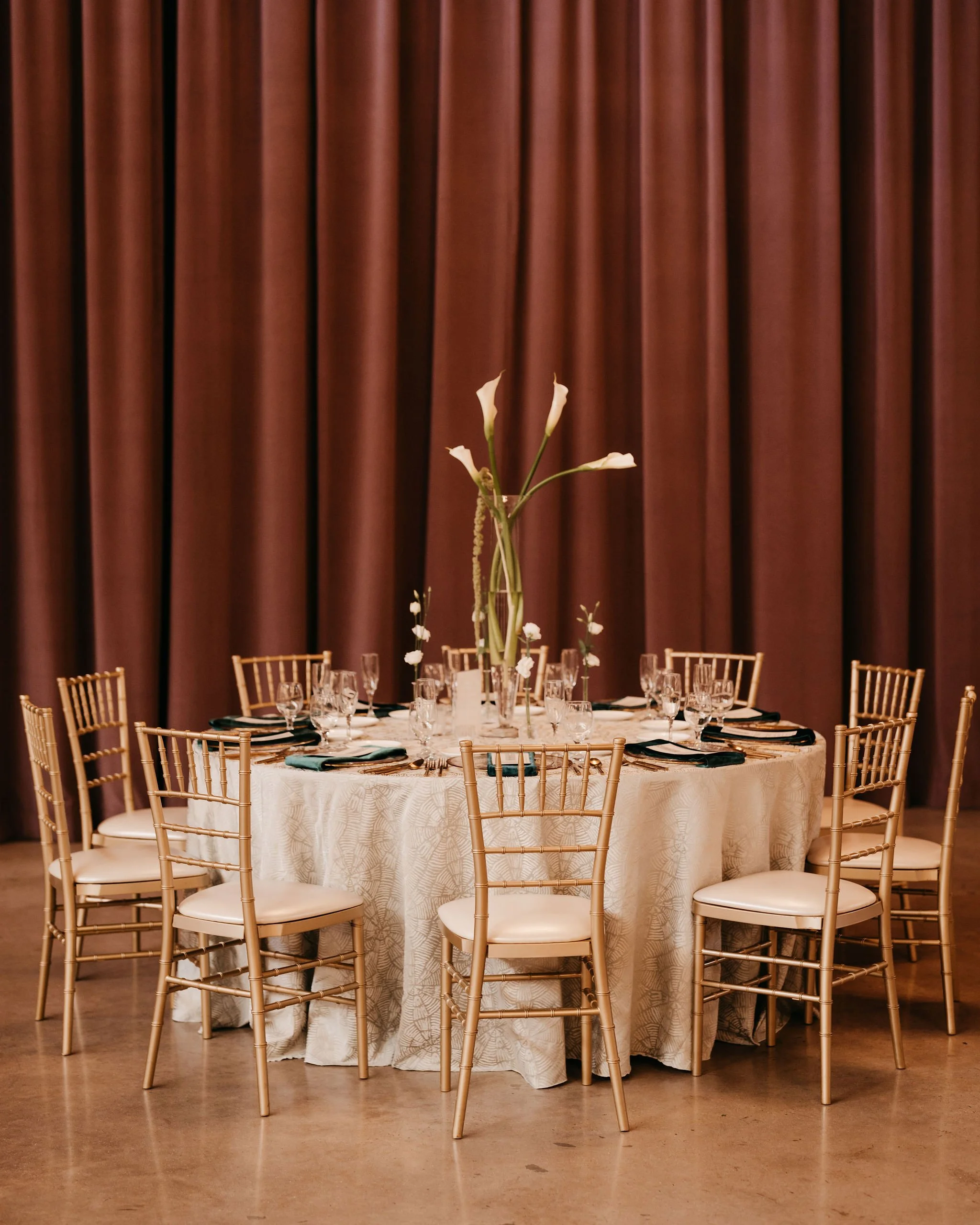 Elegant round dining table set for a formal event with tall flower centerpiece, surrounded by gold Chiavari chairs, against a backdrop of floor-length curtains.