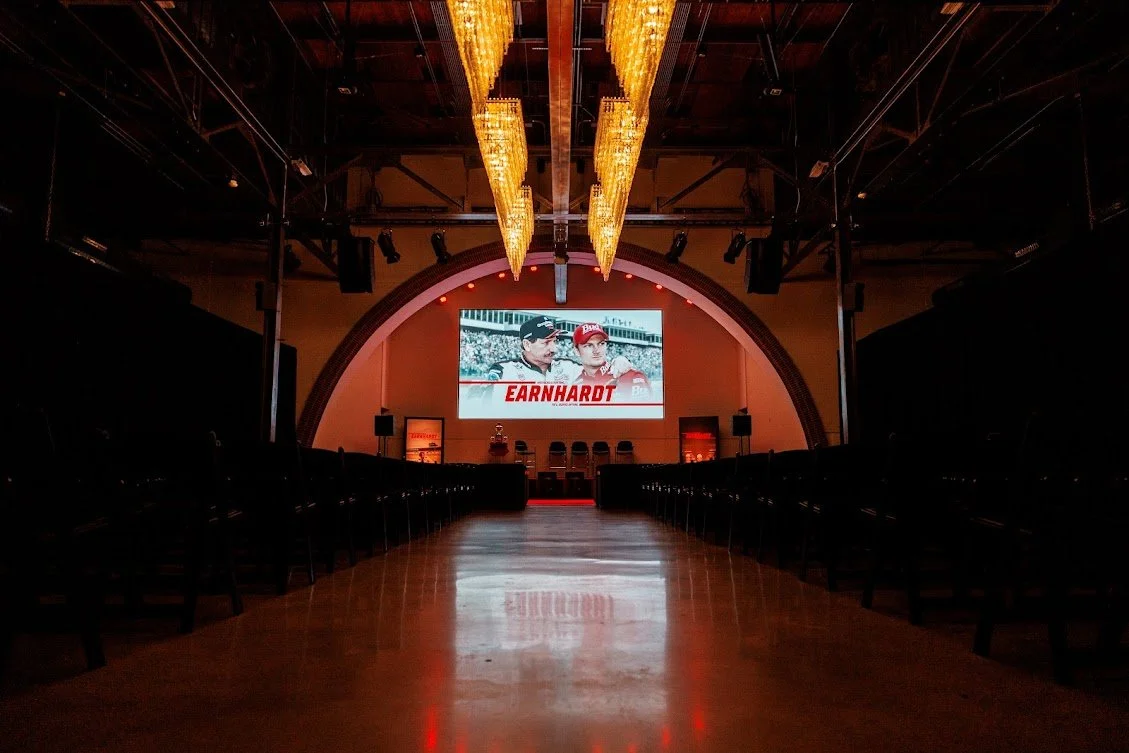 An empty auditorium with a large screen displaying two race car drivers and the name 'Earnhardt', with chairs arranged in rows on either side of the aisle, and chandeliers hanging from the ceiling.