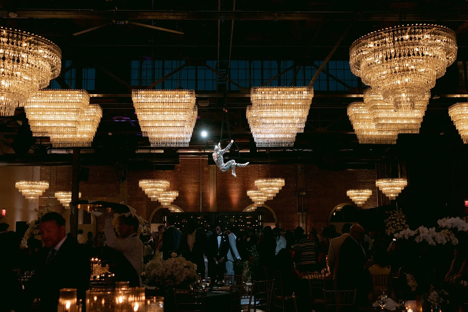 Indoor event space with multiple large chandeliers hanging from the ceiling. In the center, a performer in a shiny outfit is swinging on a trapeze or aerial hoop. The room has dark walls, some tables with candles, and many people dressed in formal at