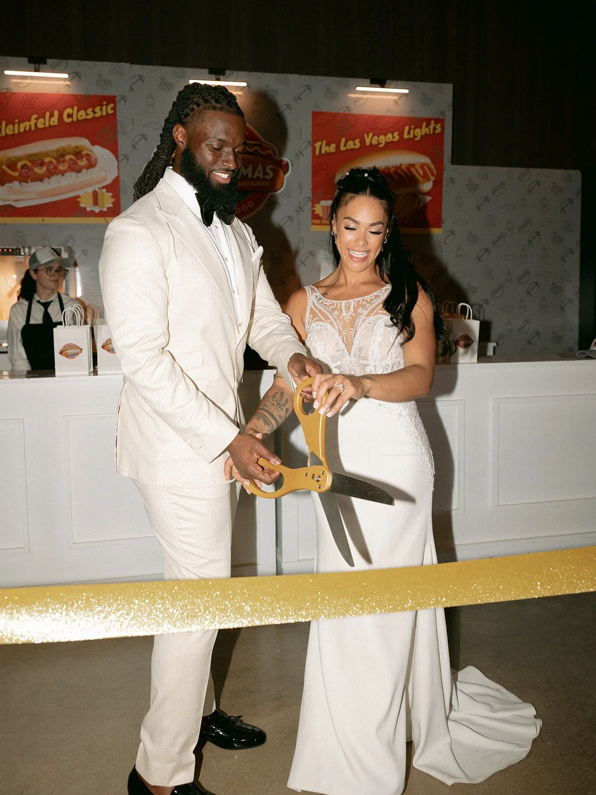 A couple in wedding attire cutting a gold ribbon with large scissors at a celebratory event.