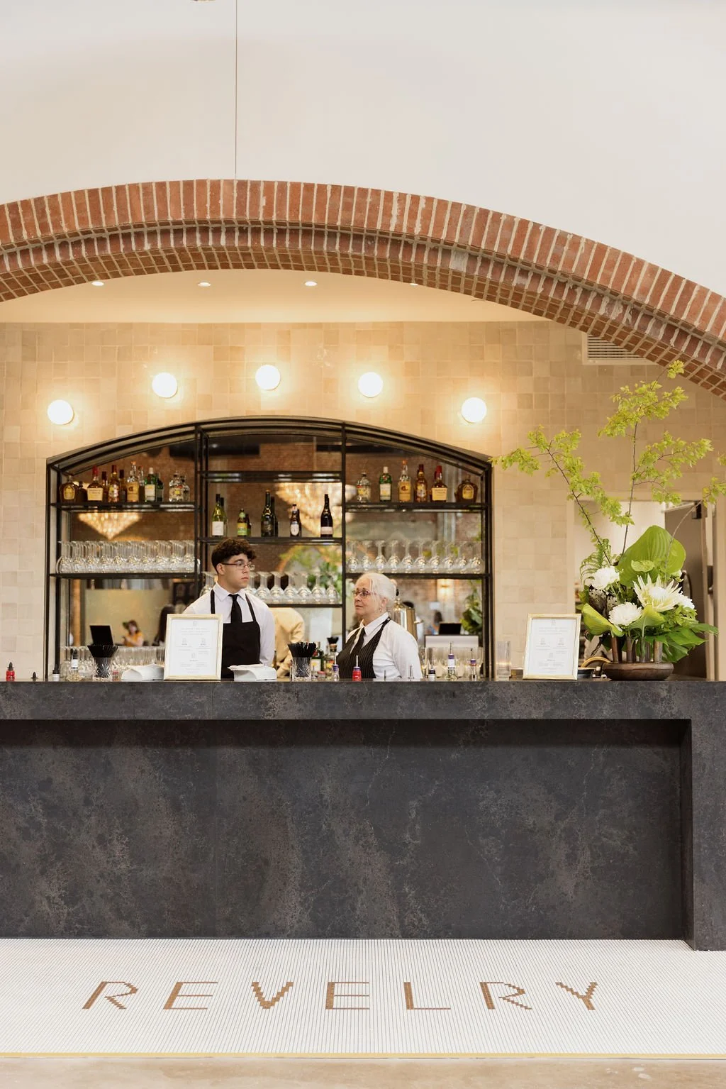 A bar counter in a restaurant with two staff members, one young man and an older woman, behind it. Shelves with bottles and glasses are in the background, with a decorative plant on the right side. The area features a brick arch and warm lighting. Th